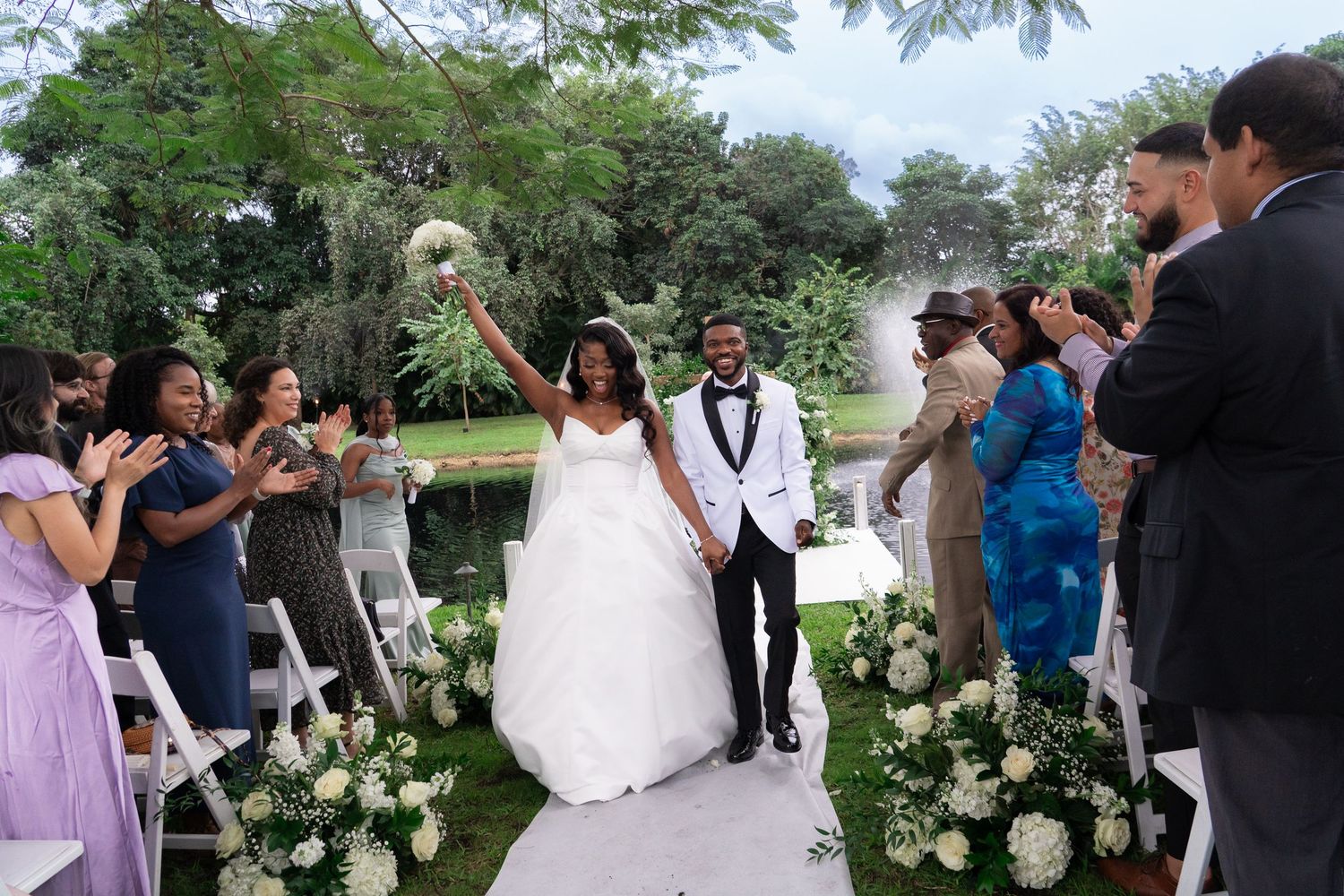 Joyful newlywed couple walks down outdoor aisle after wedding ceremony, bride raising bouquet, guests applauding on both sides.
