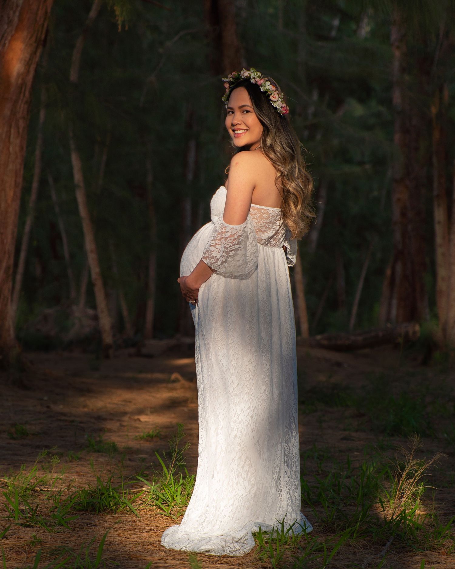 A smiling pregnant woman in a white lace off-shoulder gown wearing a floral crown poses in a forest.