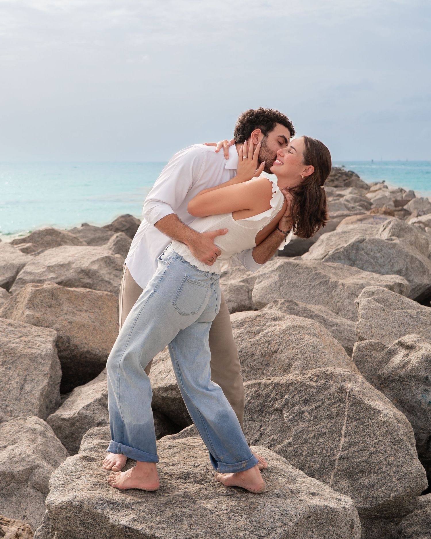 Couple kissing romantically on rocky beach shoreline, man dipping woman in white top and jeans by turquoise ocean.