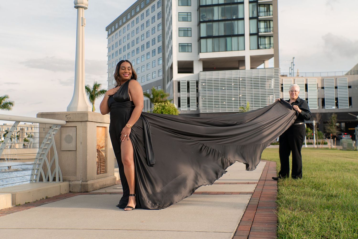 Woman in a dramatic black gown with a flowing train posing outdoors near a city waterfront.