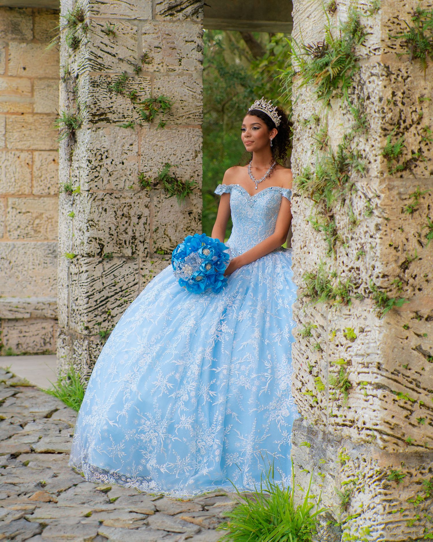 Elegant young woman in a light blue lace quinceañera gown holding a blue bouquet near stone columns.