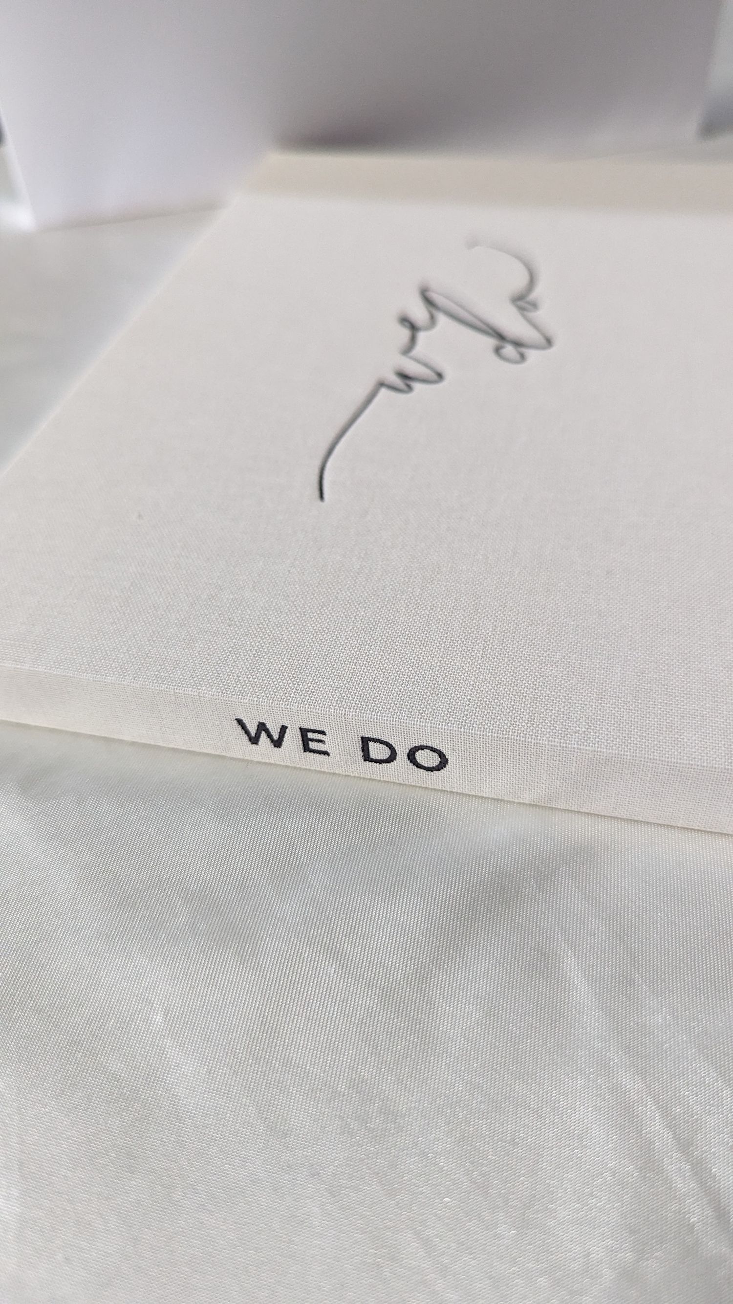 Elegant white linen wedding guest book with cursive design and WE DO text on cover.