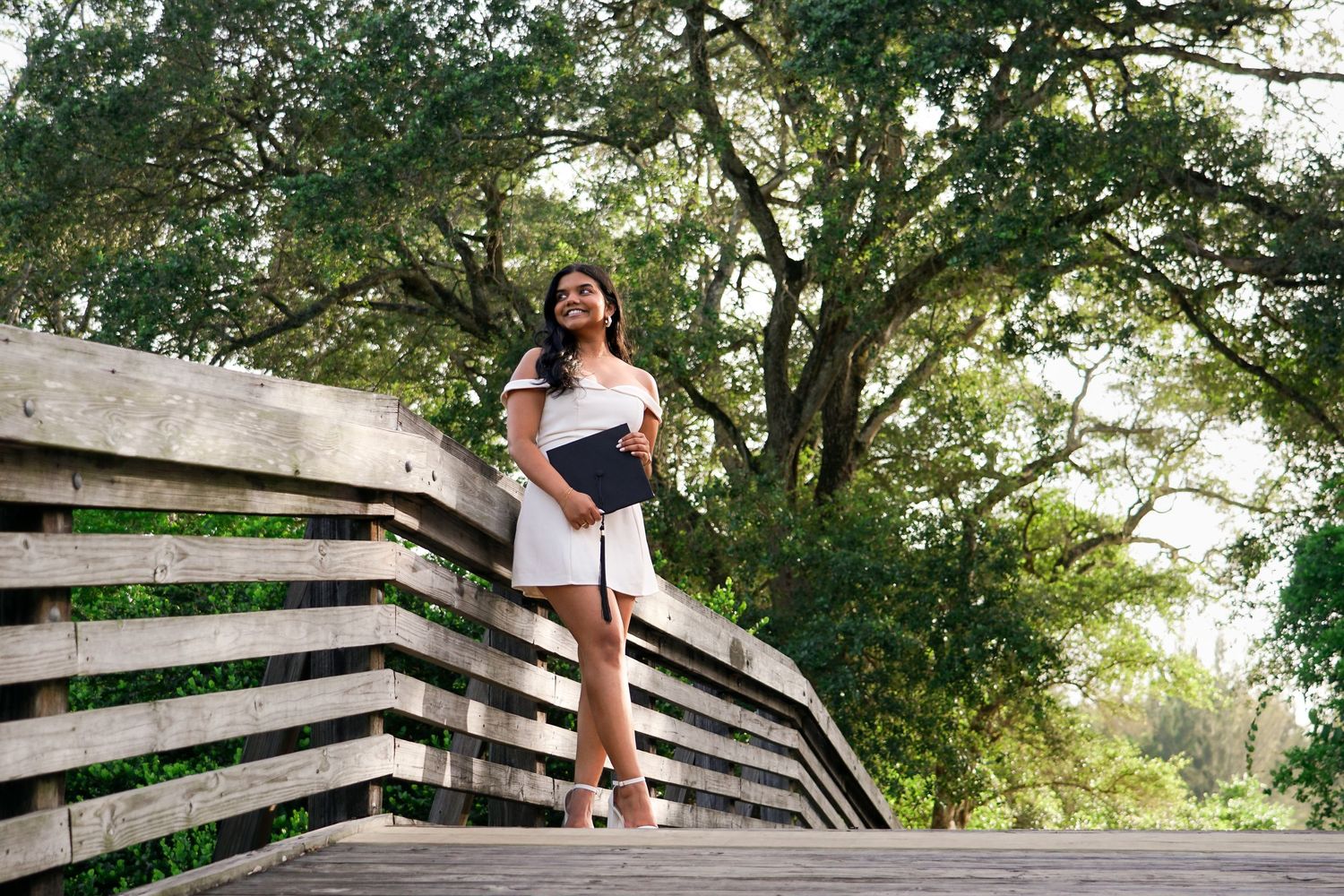 Young woman in a white dress holding a diploma on a wooden bridge surrounded by lush greenery.