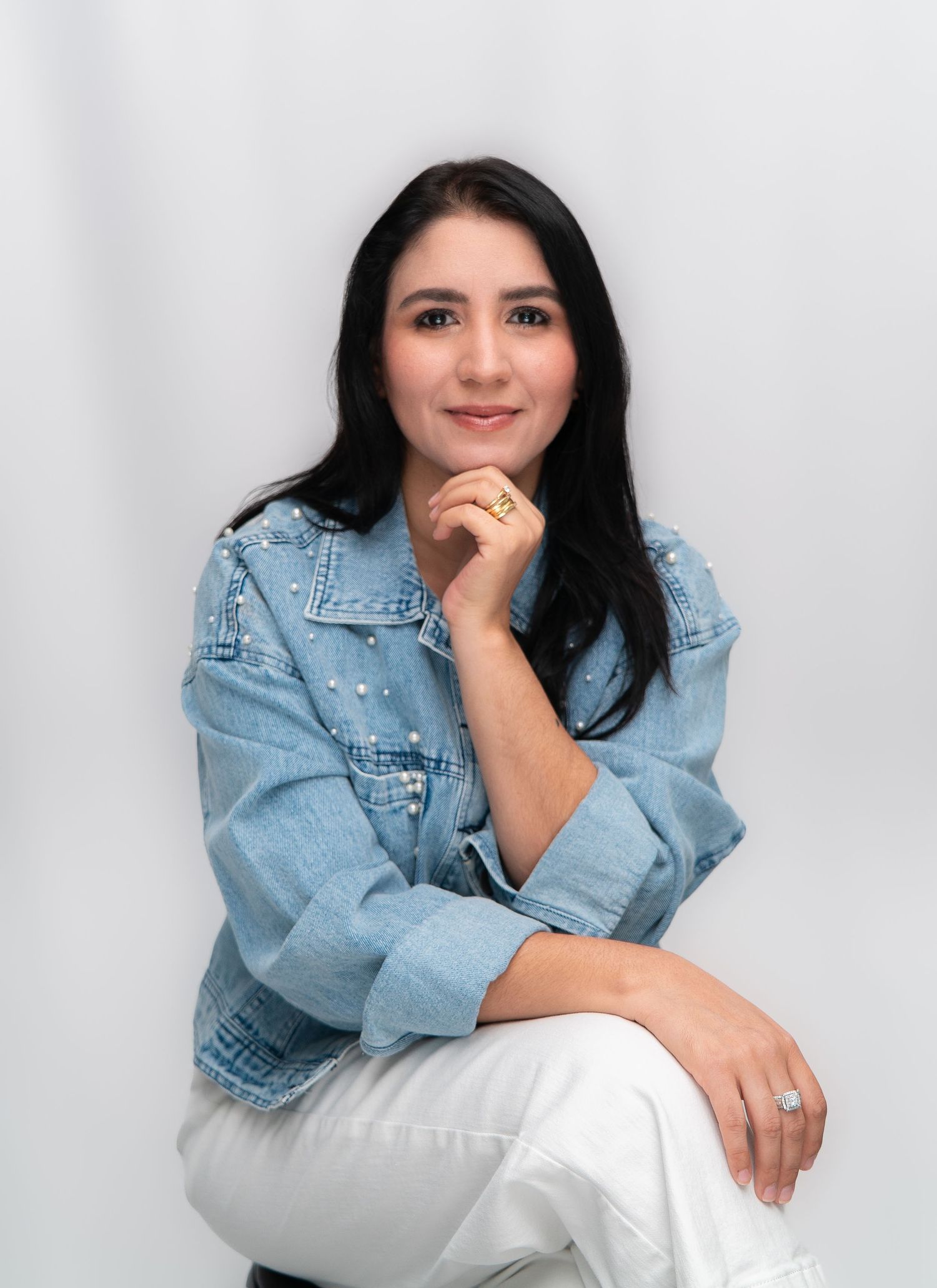 A young woman in a denim jacket sits gracefully with her hand on her chin against a white background.