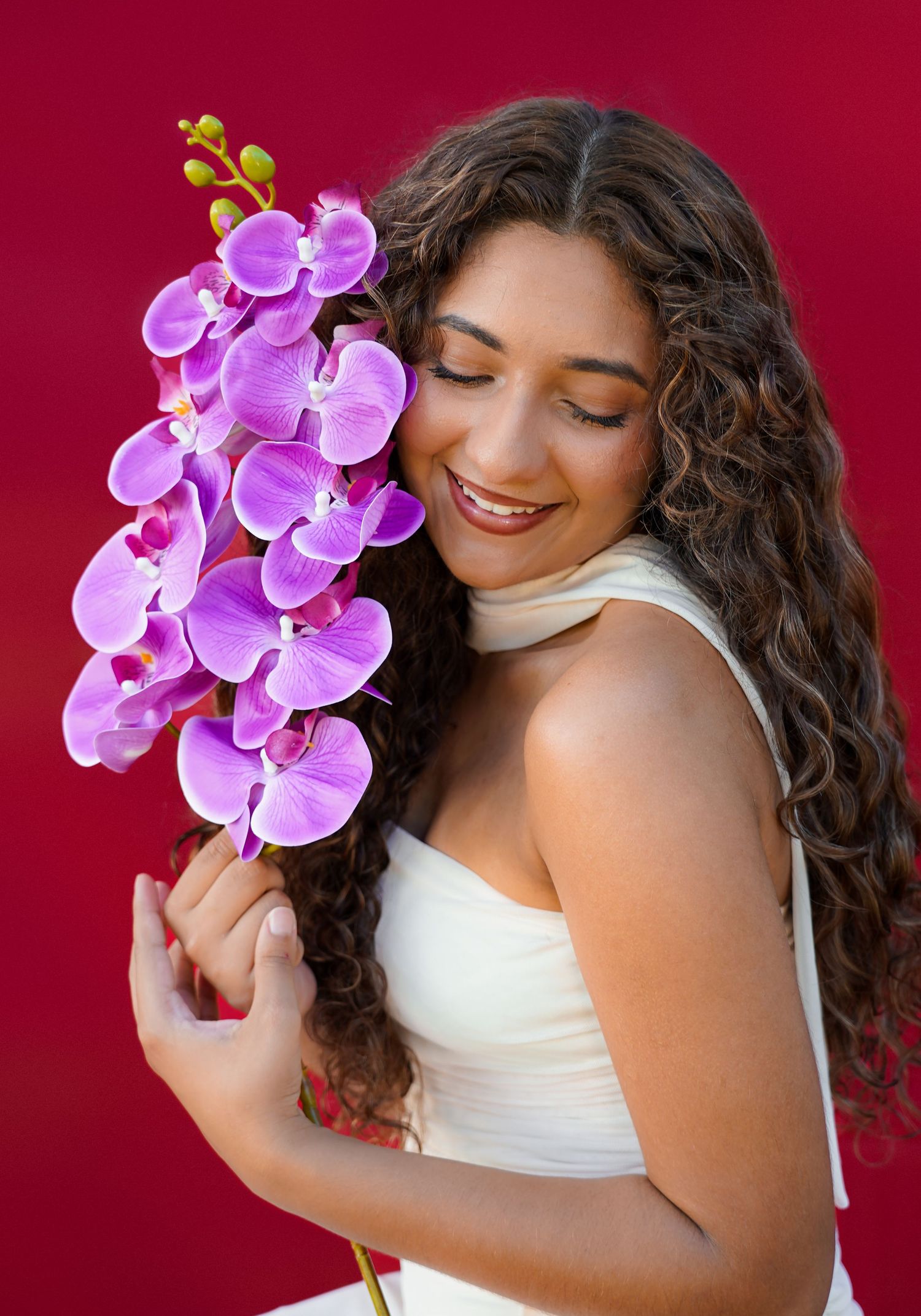 A smiling woman with curly hair holds purple orchid flowers against a vibrant red background.