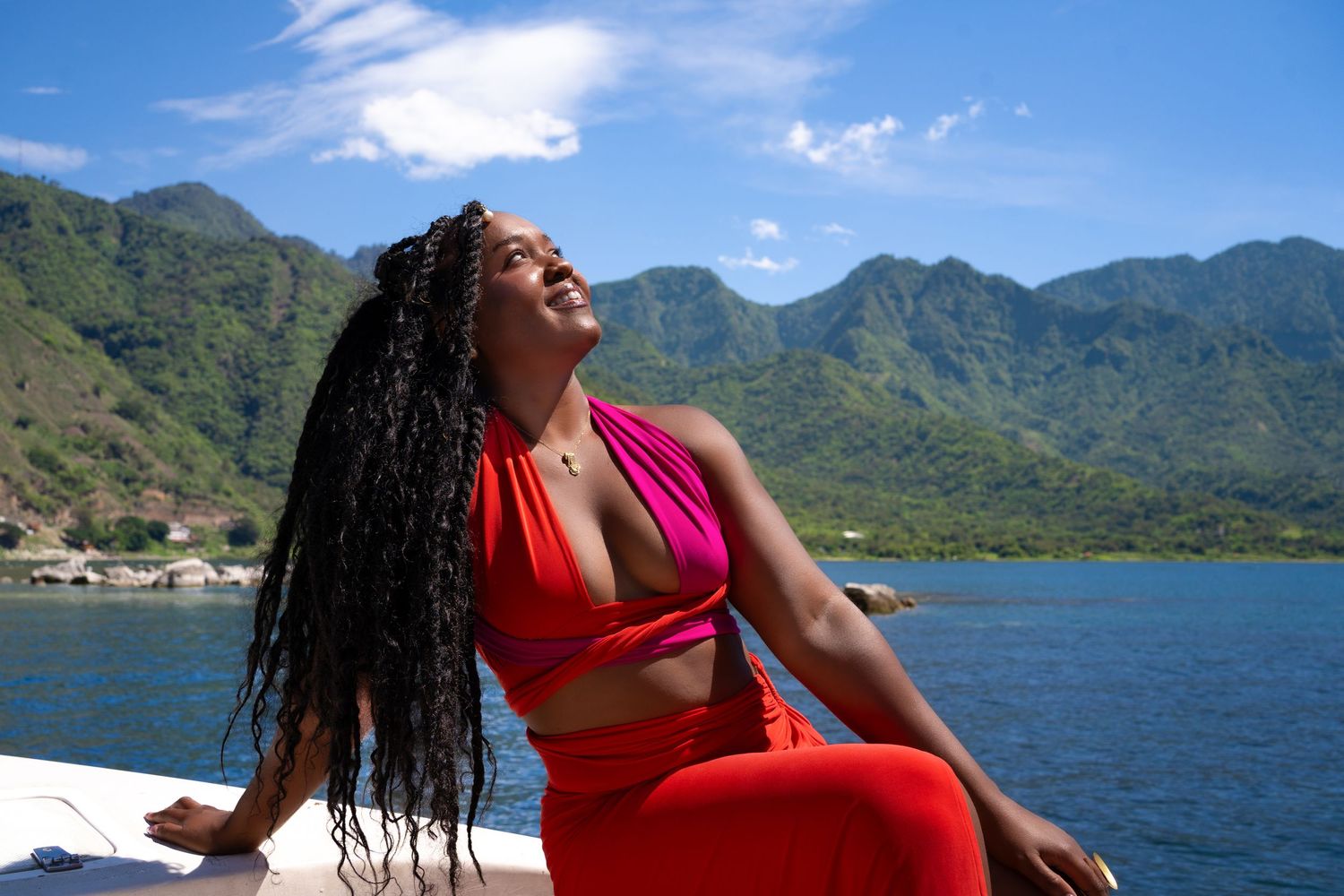 Woman in red outfit relaxing by tropical mountain lake under blue sky with lush green hills in background.