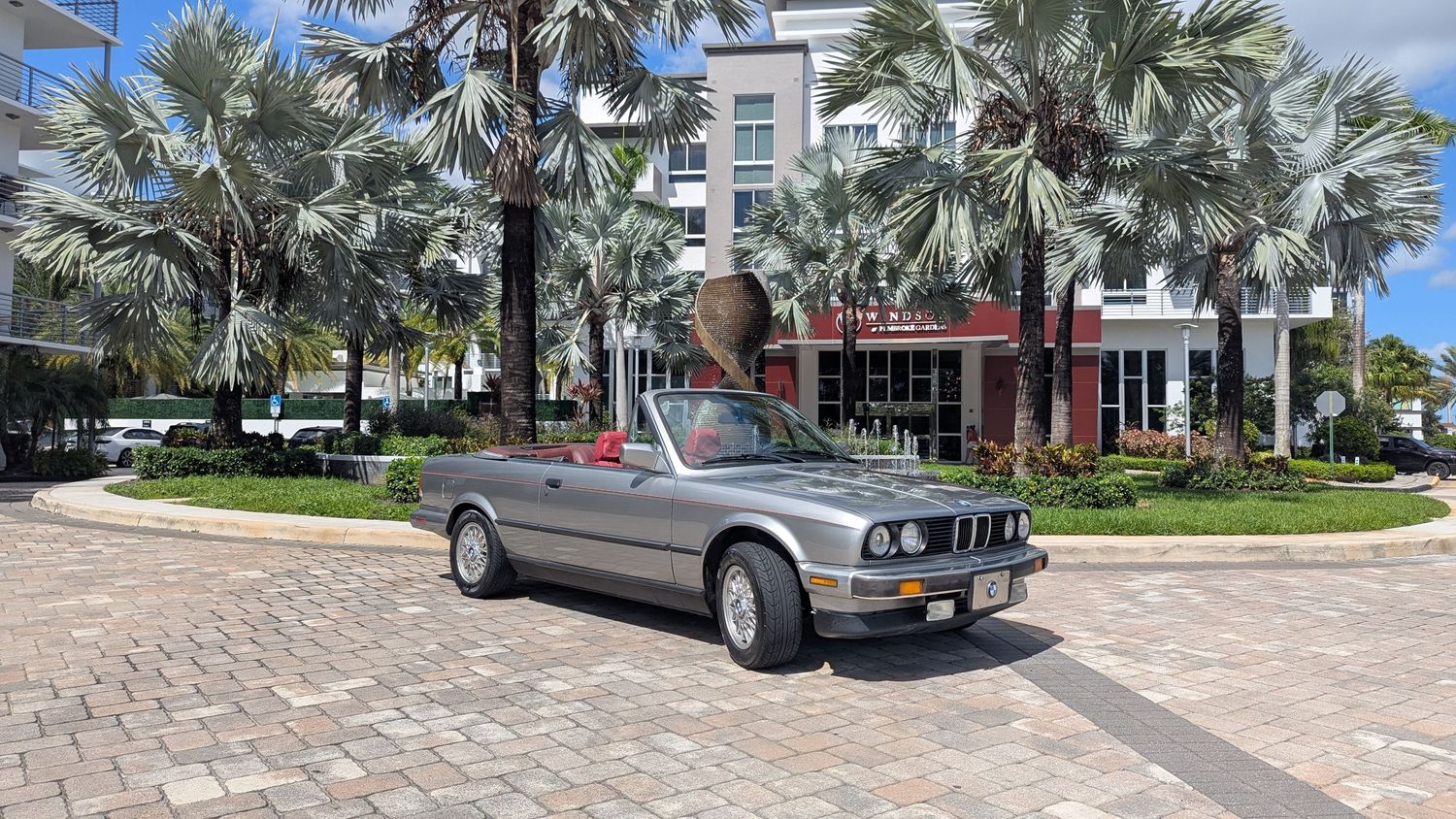 A silver BMW E30 convertible parked on brick pavers surrounded by palm trees near a modern building on a sunny day.