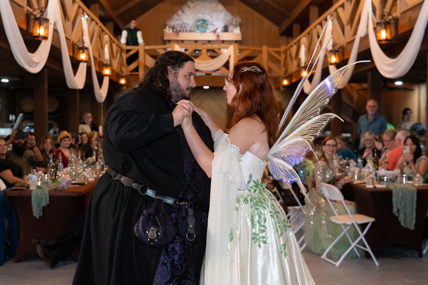 Newlyweds dancing at fairy tale wedding reception, bride wearing iridescent wings and leaf-embroidered white gown.