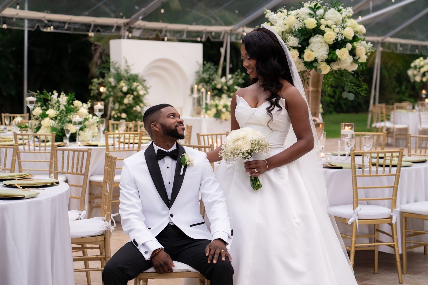 Bride holding bouquet stands beside seated groom at outdoor tented wedding reception.