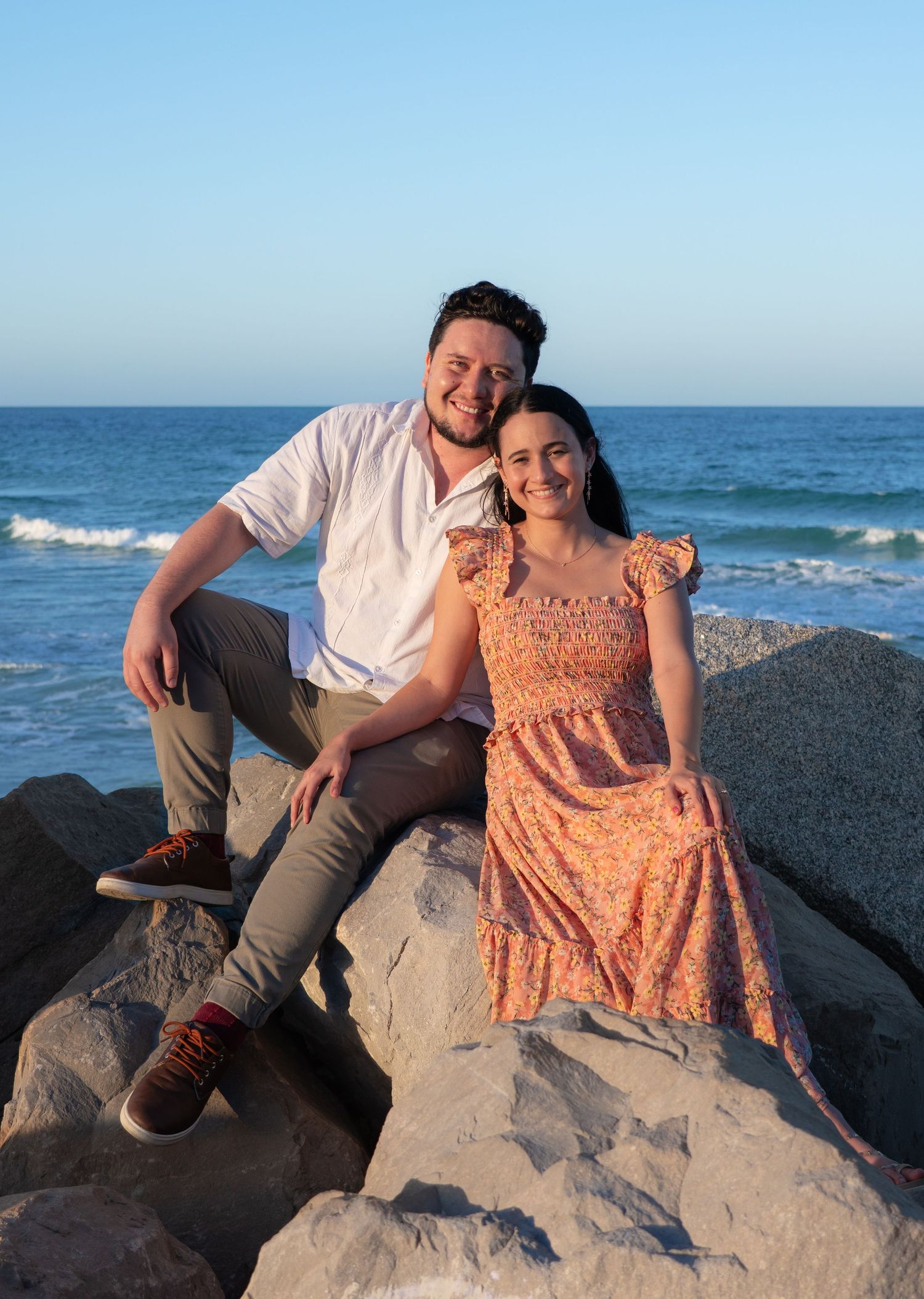 Couple smiling and sitting on rocks at the beach with ocean waves and blue sky in the background.
