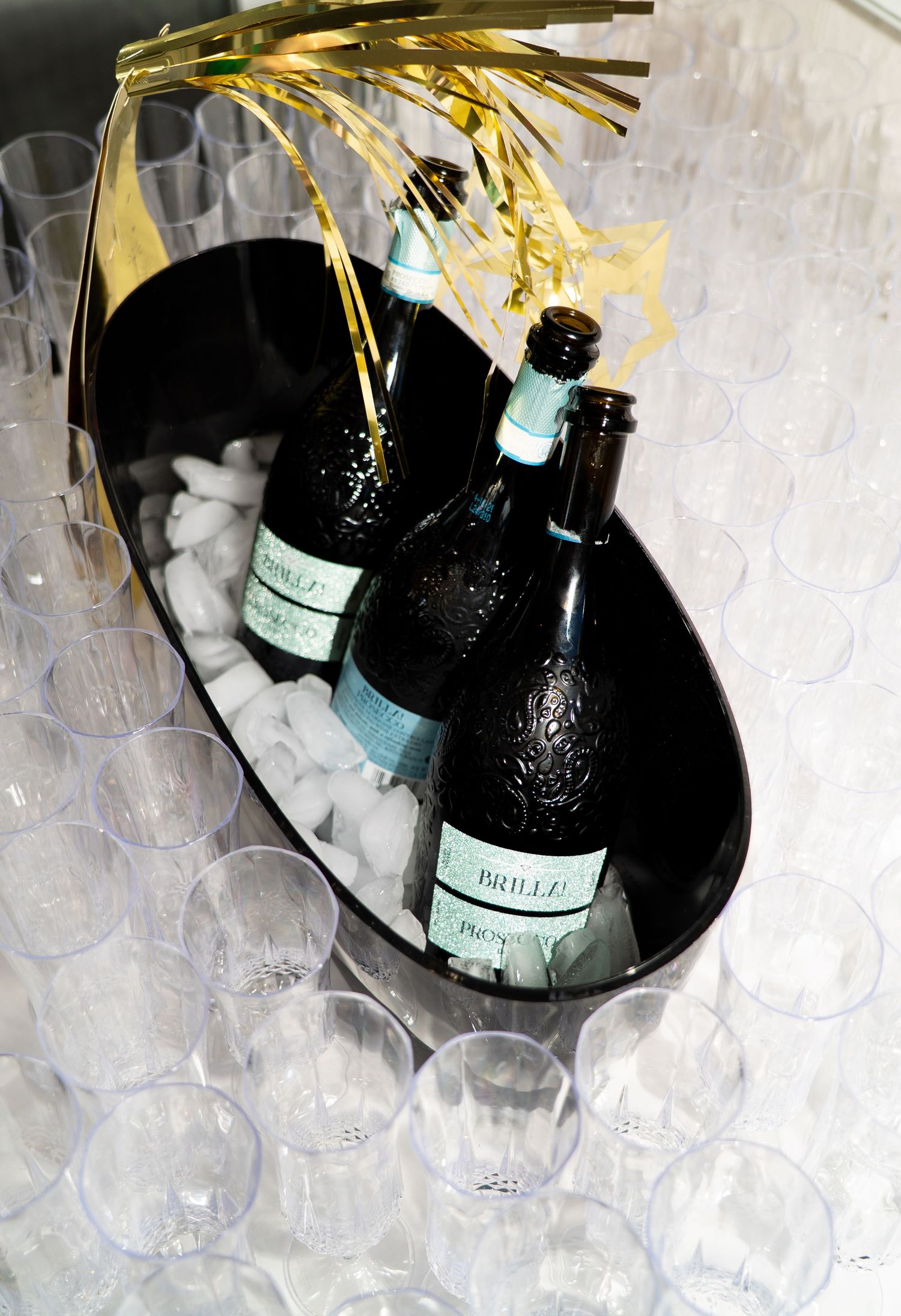 Champagne bottles chilling on ice in a black oval bucket surrounded by clear plastic glasses at a party.