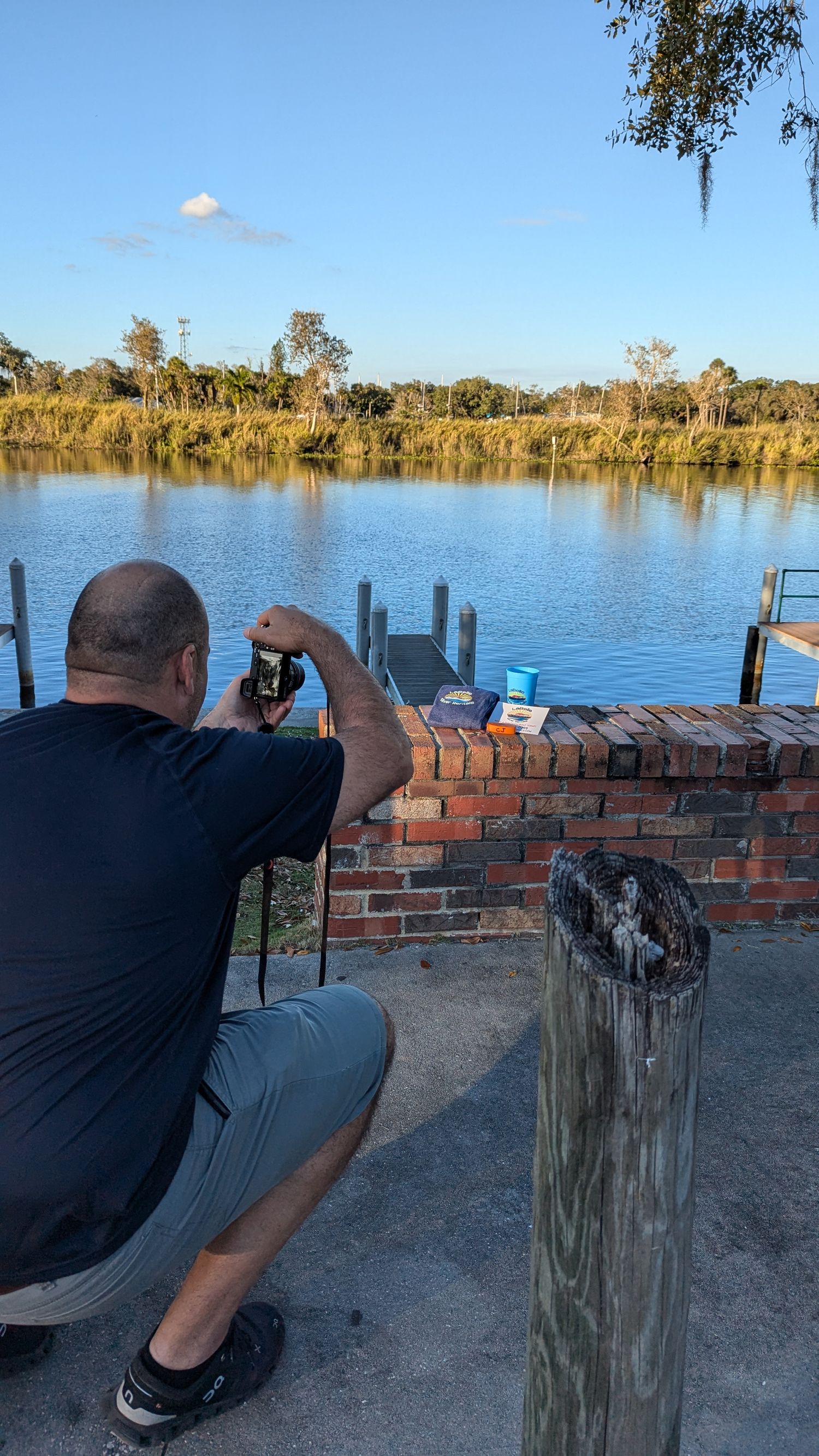 Man crouching by brick wall photographing a serene Florida waterway with dock and lush greenery on a sunny day.
