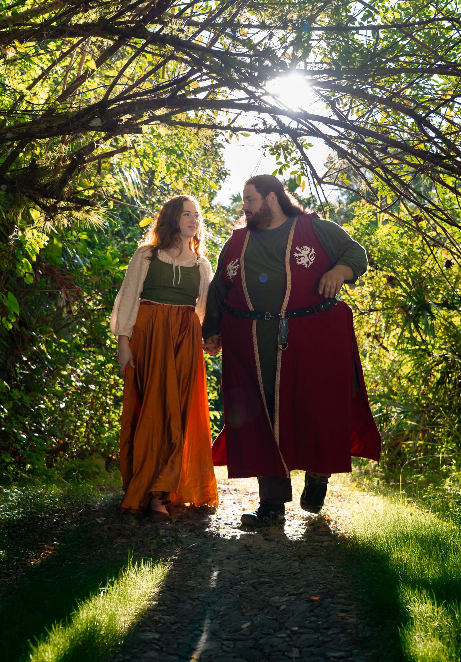 Couple in medieval Renaissance costumes walking hand in hand through a sunlit garden archway.