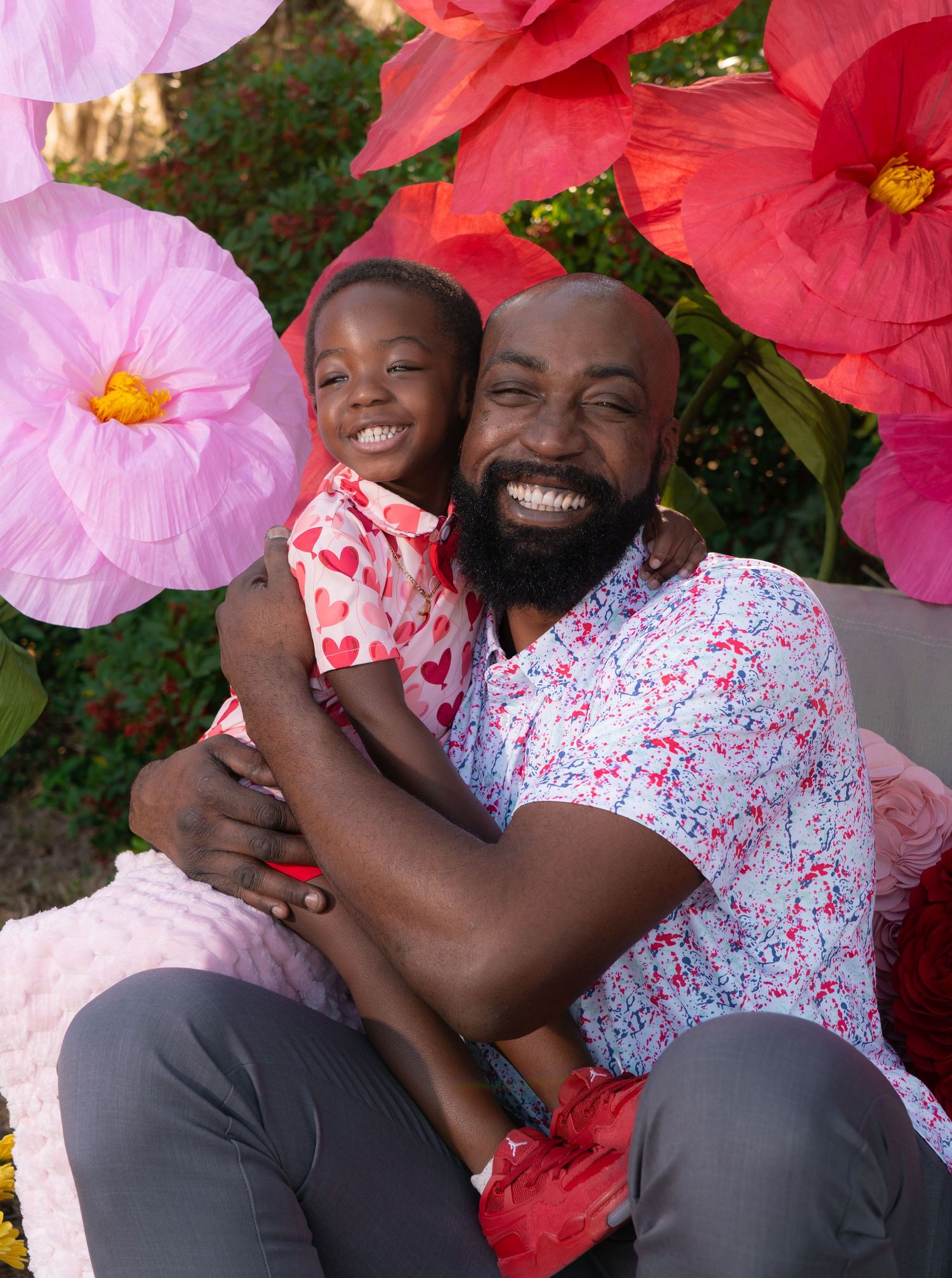 Smiling father with beard hugging young daughter surrounded by colorful paper flowers outdoors.