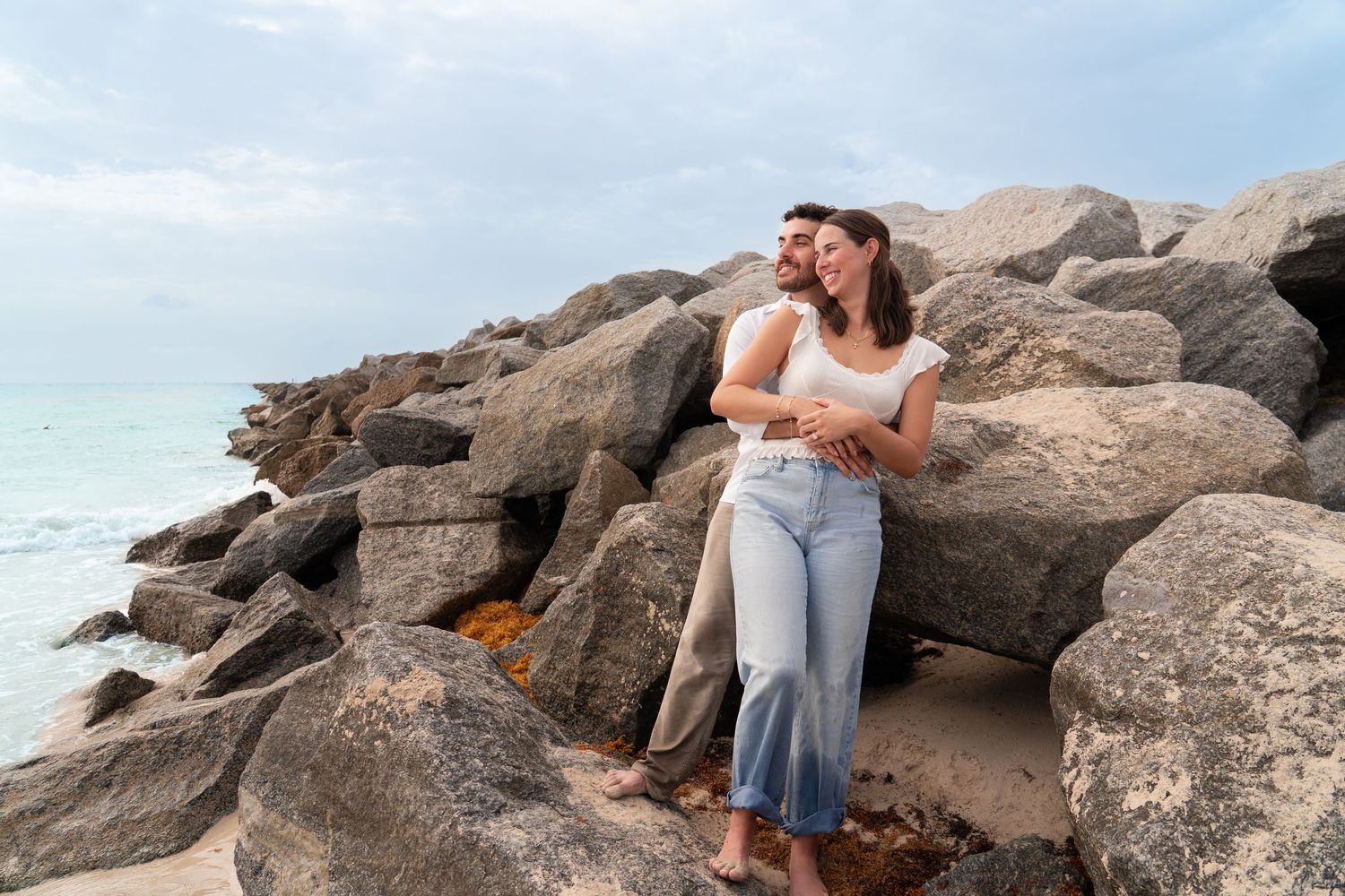 Couple embracing on rocky beach shore with turquoise ocean waves in background on cloudy day.