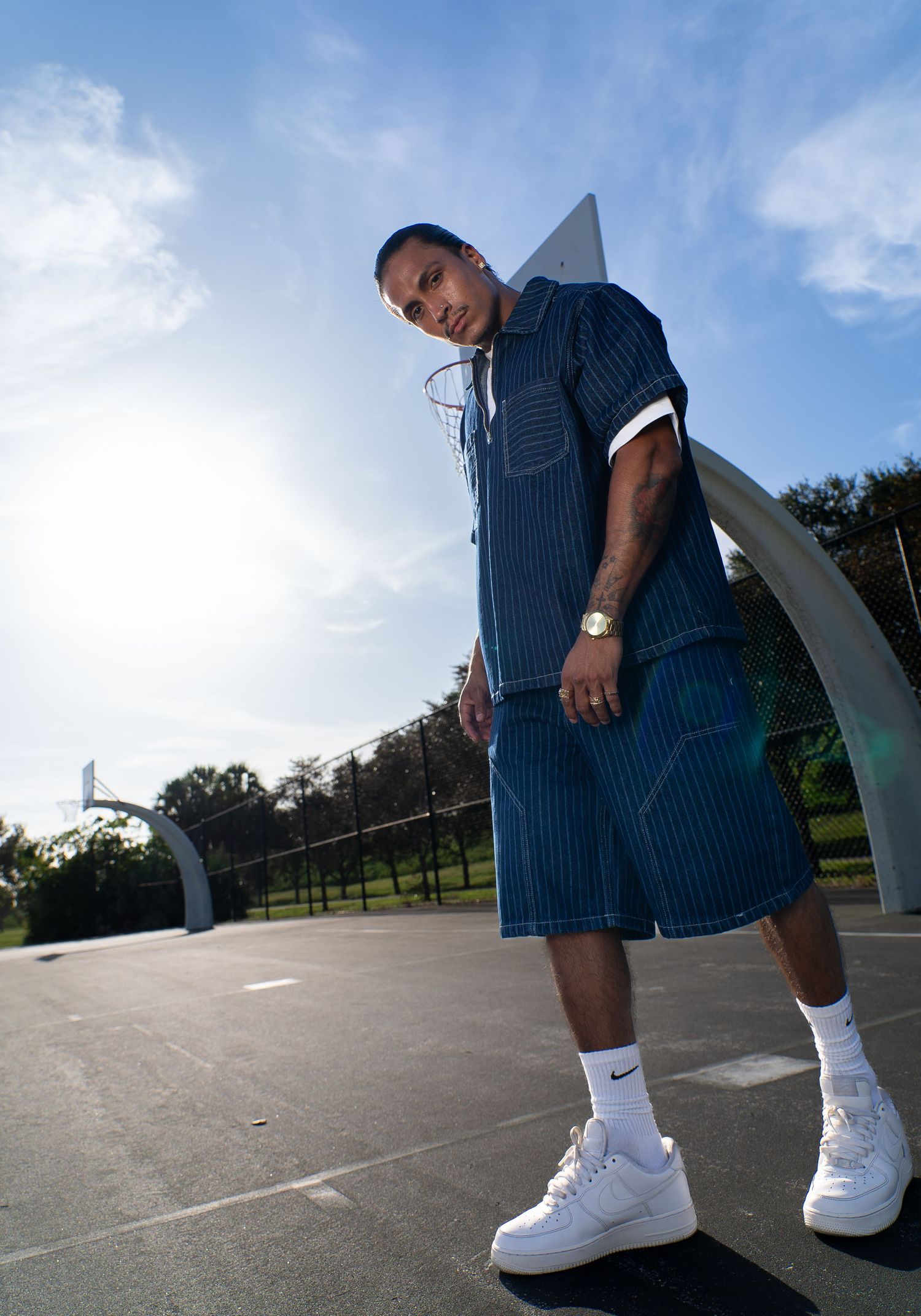Man in blue striped outfit and white Nike Air Force 1s posing on outdoor basketball court under blue sky.