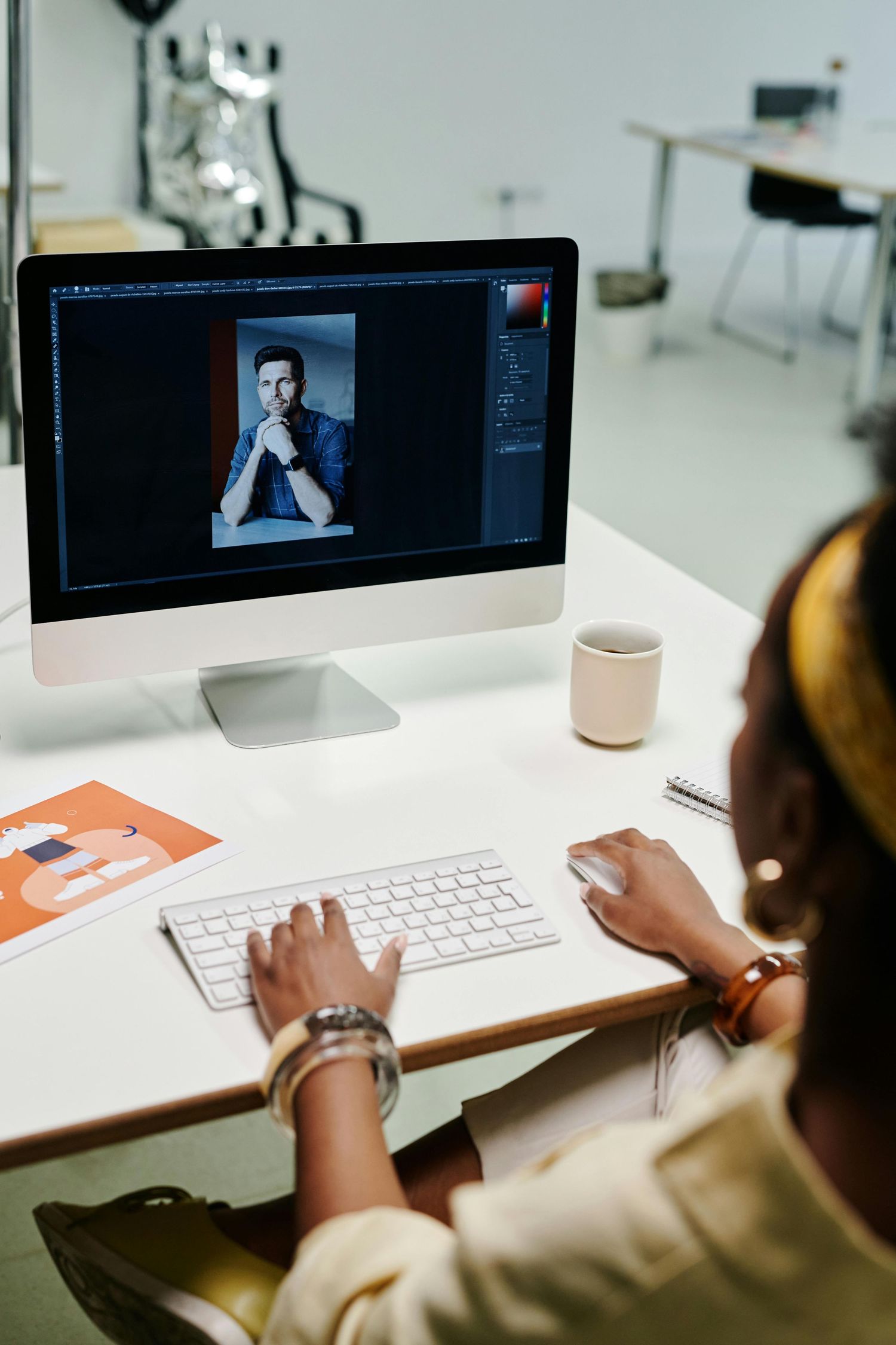 A graphic designer edits a professional portrait photo in Adobe Photoshop on an iMac in a modern studio.