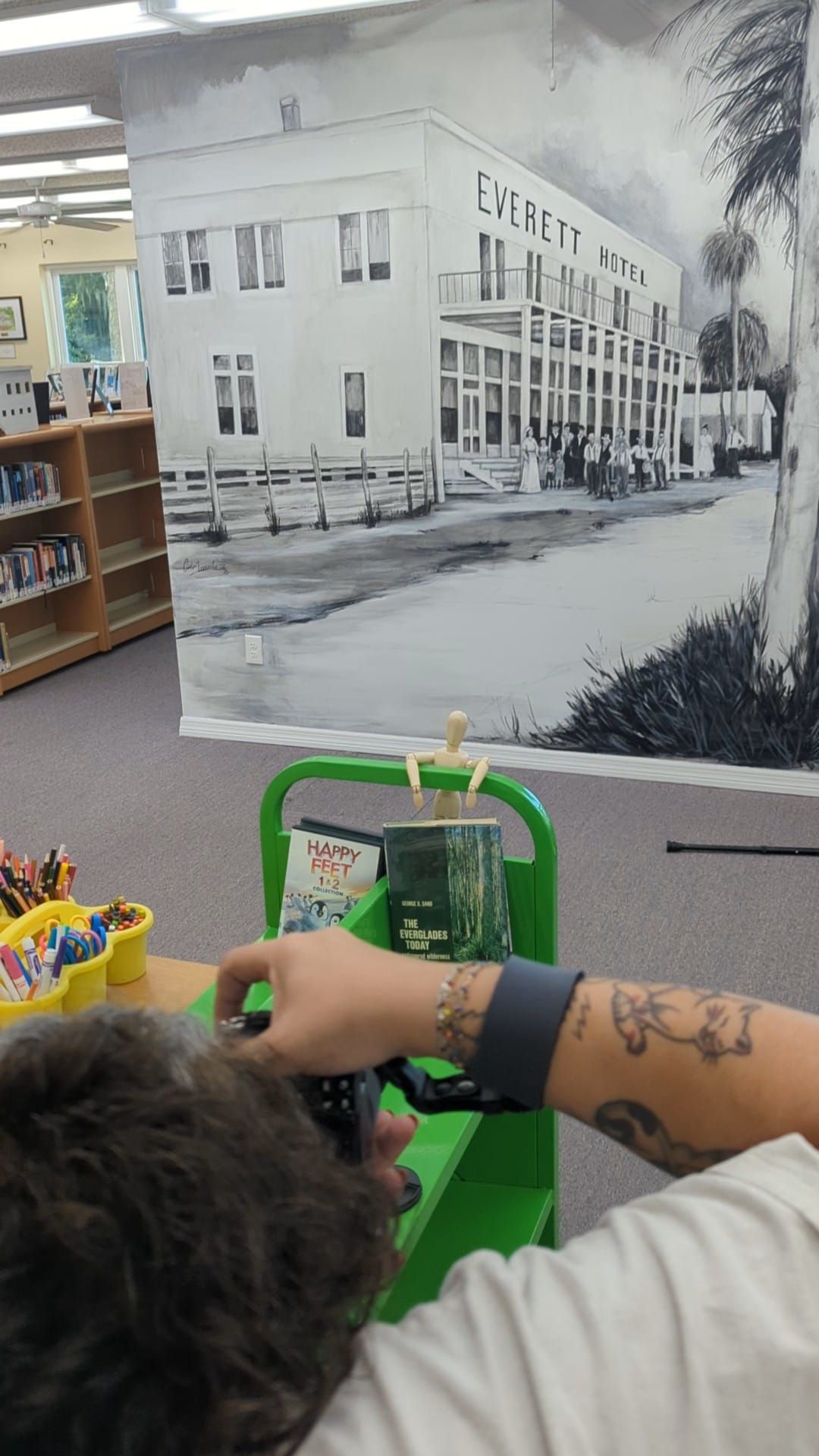 Person with tattoos browsing books at a library with a black and white Everett Hotel mural on the wall.