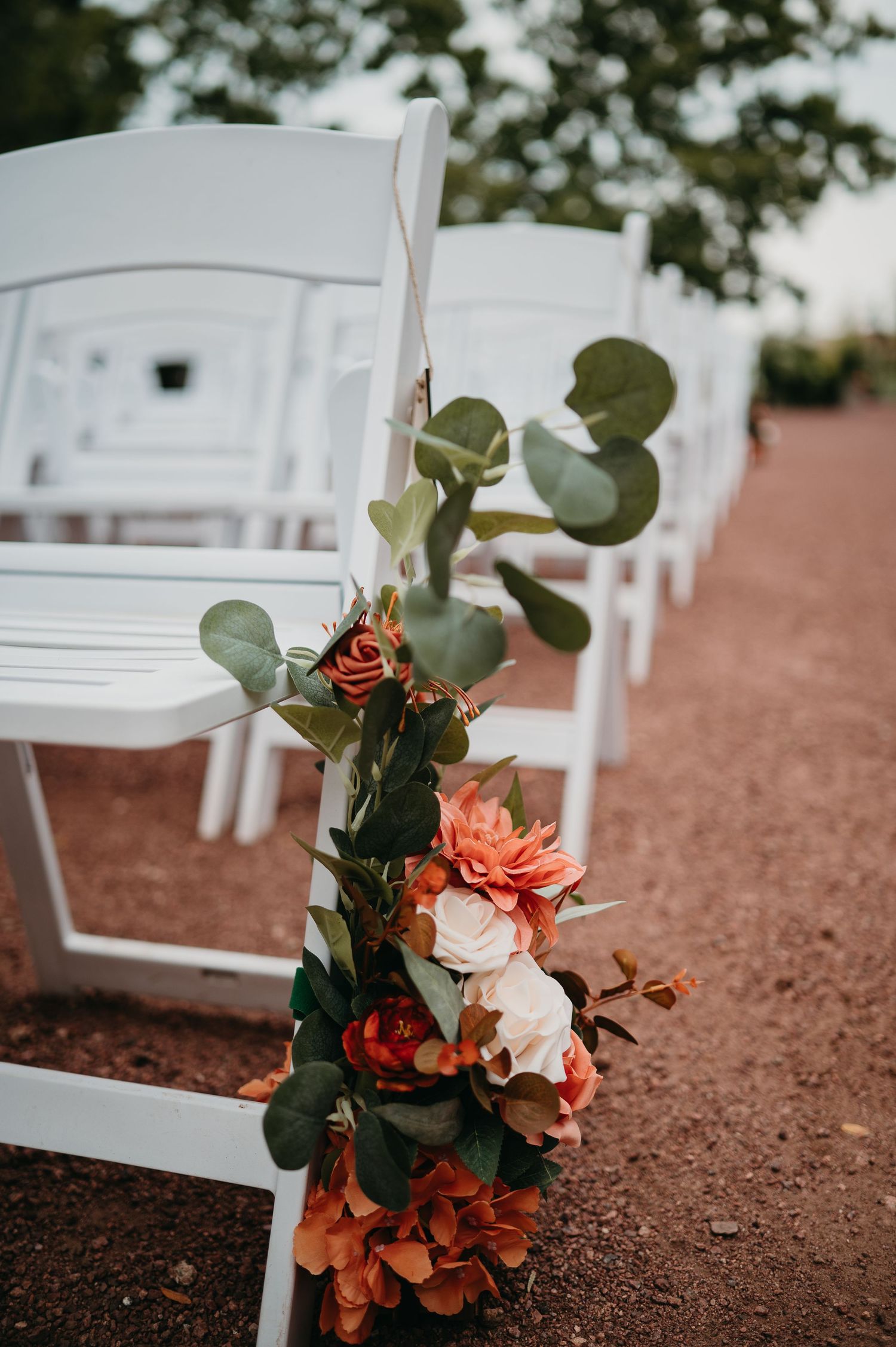 White wedding chairs arranged on red soil with coral and peach floral arrangements decorating the aisle for an outdoor ceremony.