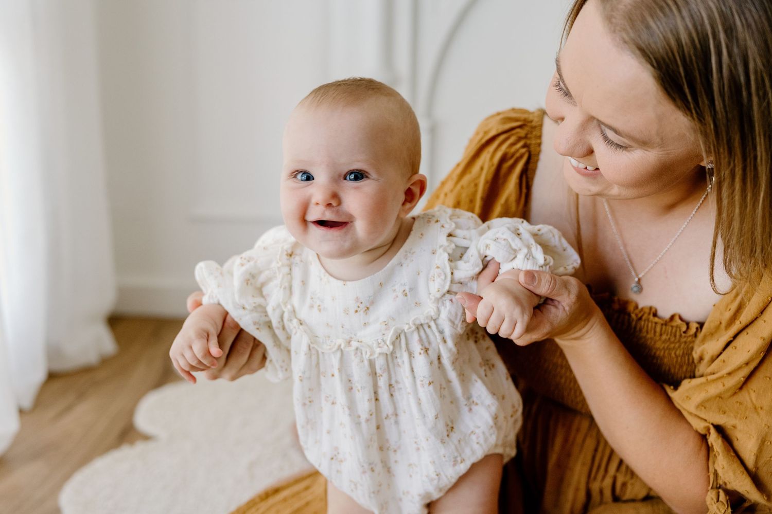 Baby wearing white lace dress smiles while being held in a bright sunny room with wooden floors.