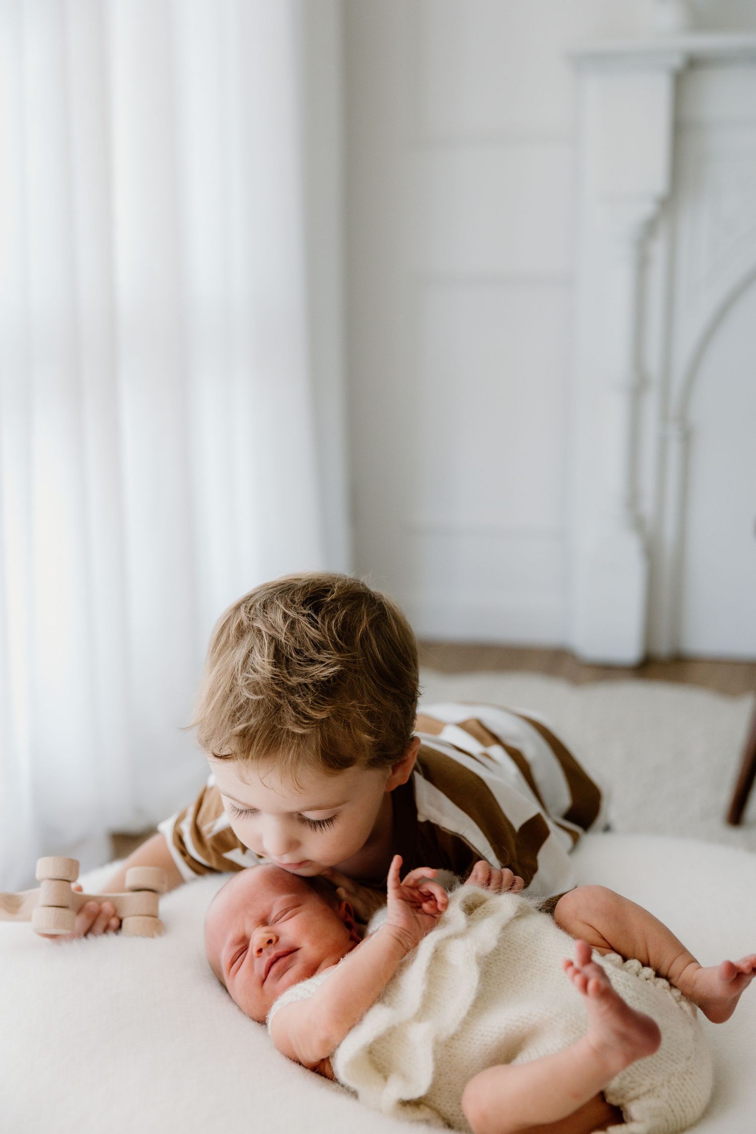Two young siblings share a tender moment on a white bed as the older one gently kisses the newborn baby.