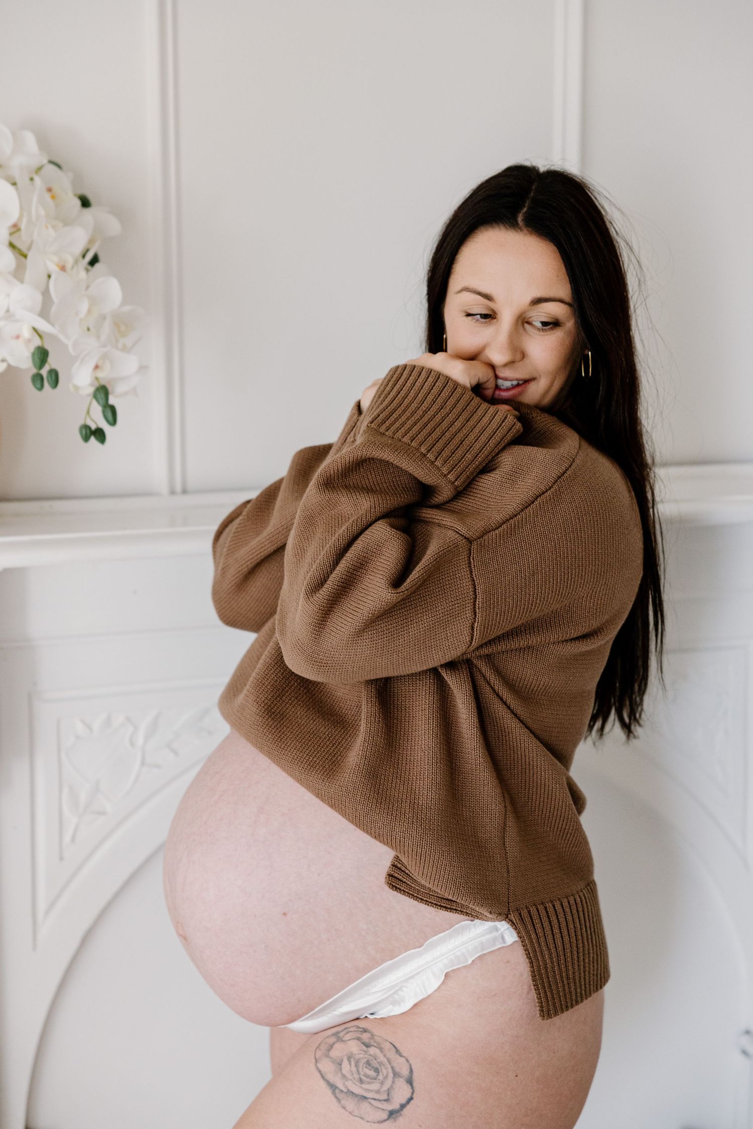 A pregnant woman in a brown sweater poses against a white wall with delicate white floral decorations.