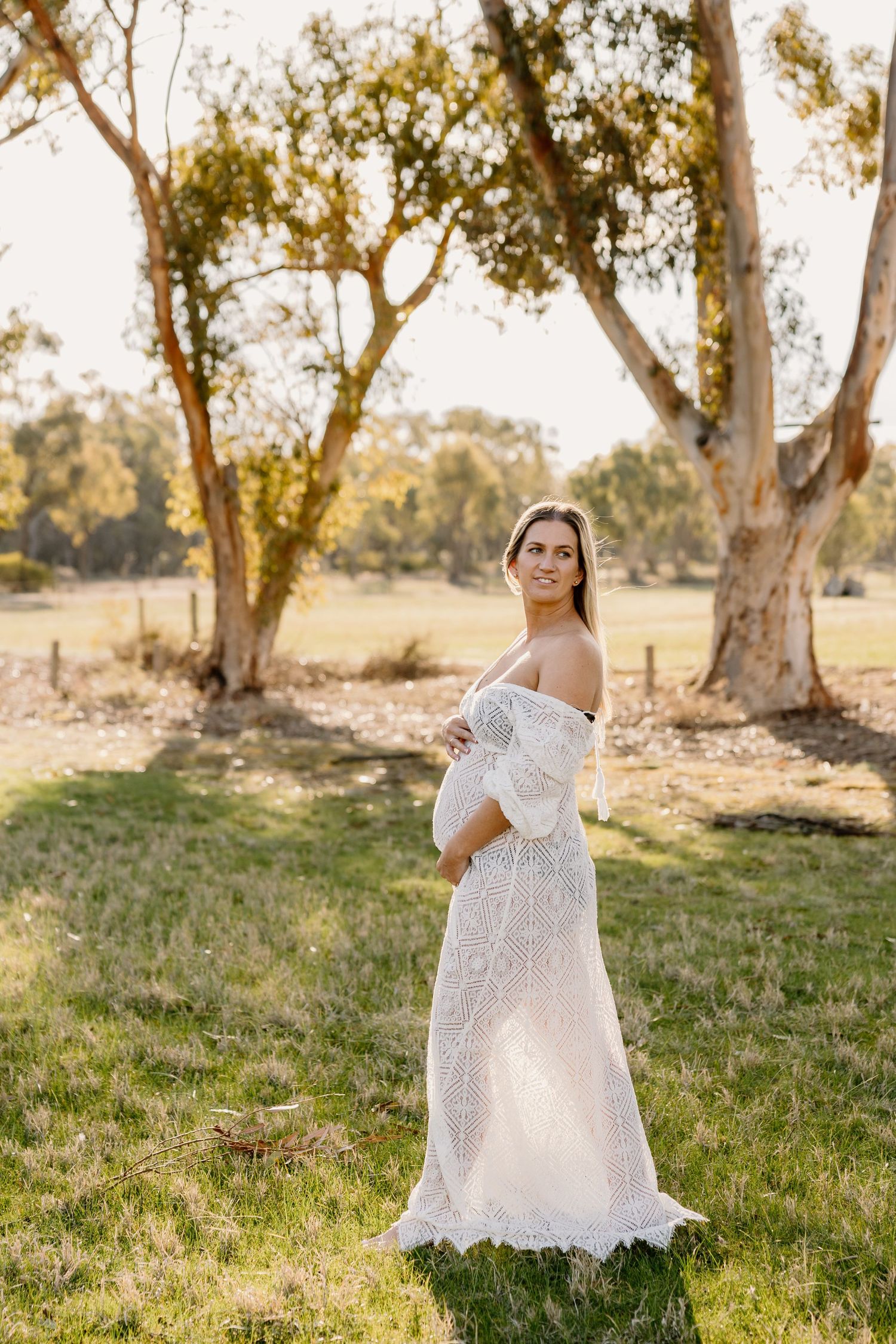 Expectant figure in flowing white lace dress poses in sunlit field with eucalyptus trees during golden hour.