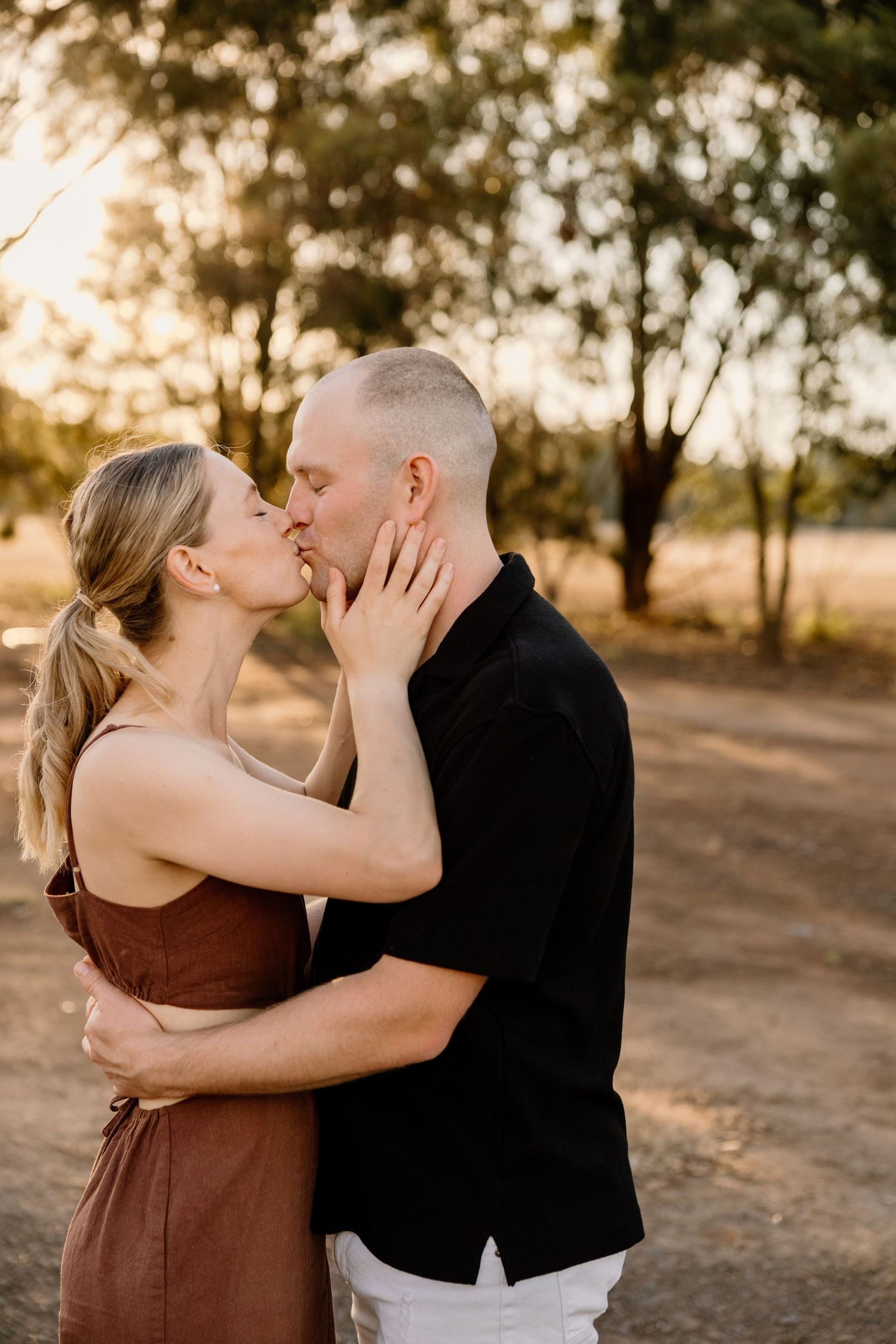 A romantic couple shares a tender kiss on a dirt path at sunset with golden sunlight filtering through trees in the background.
