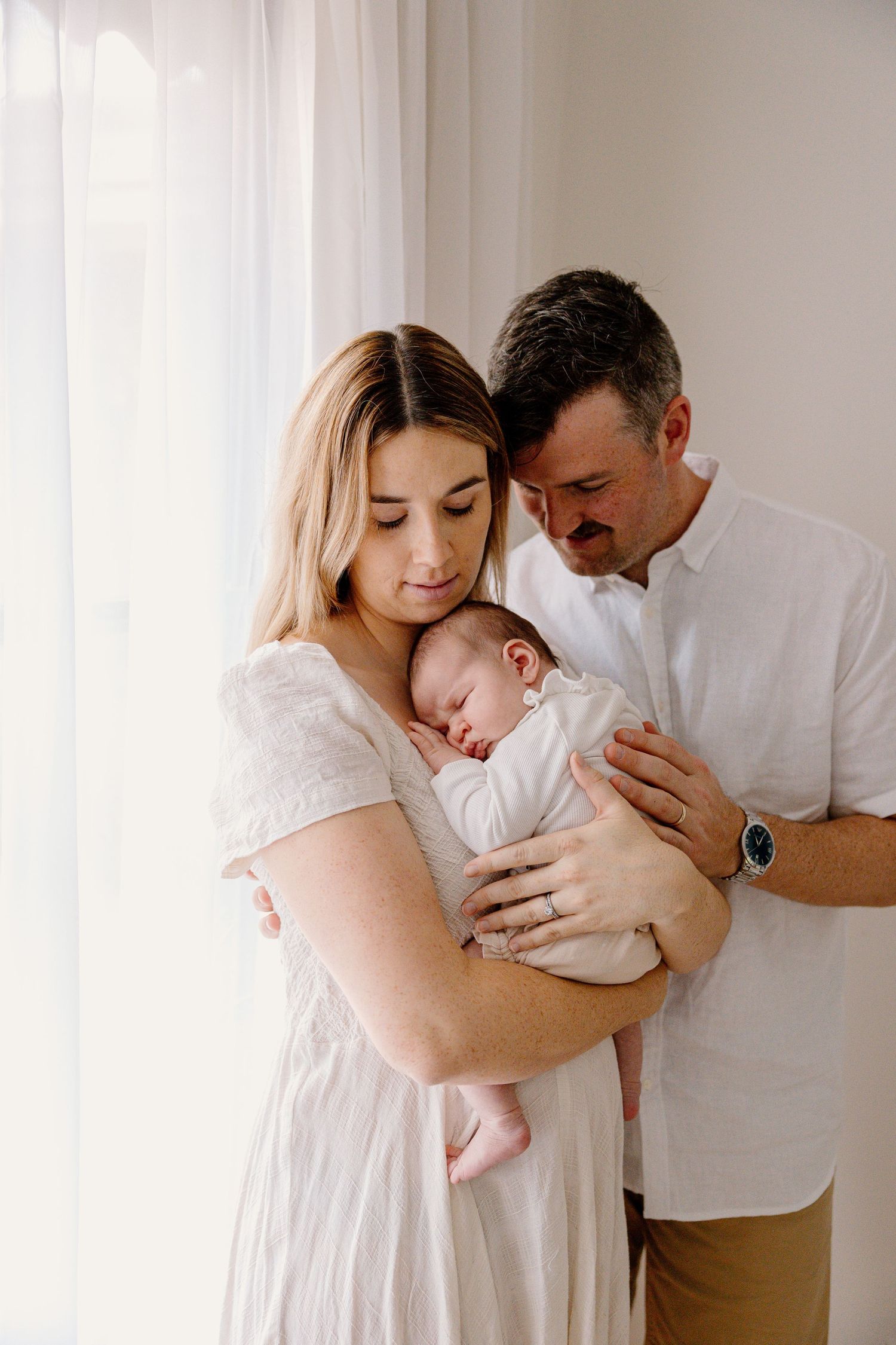 Parents in white and beige clothing gently hold their sleeping newborn baby near a window with soft natural light.