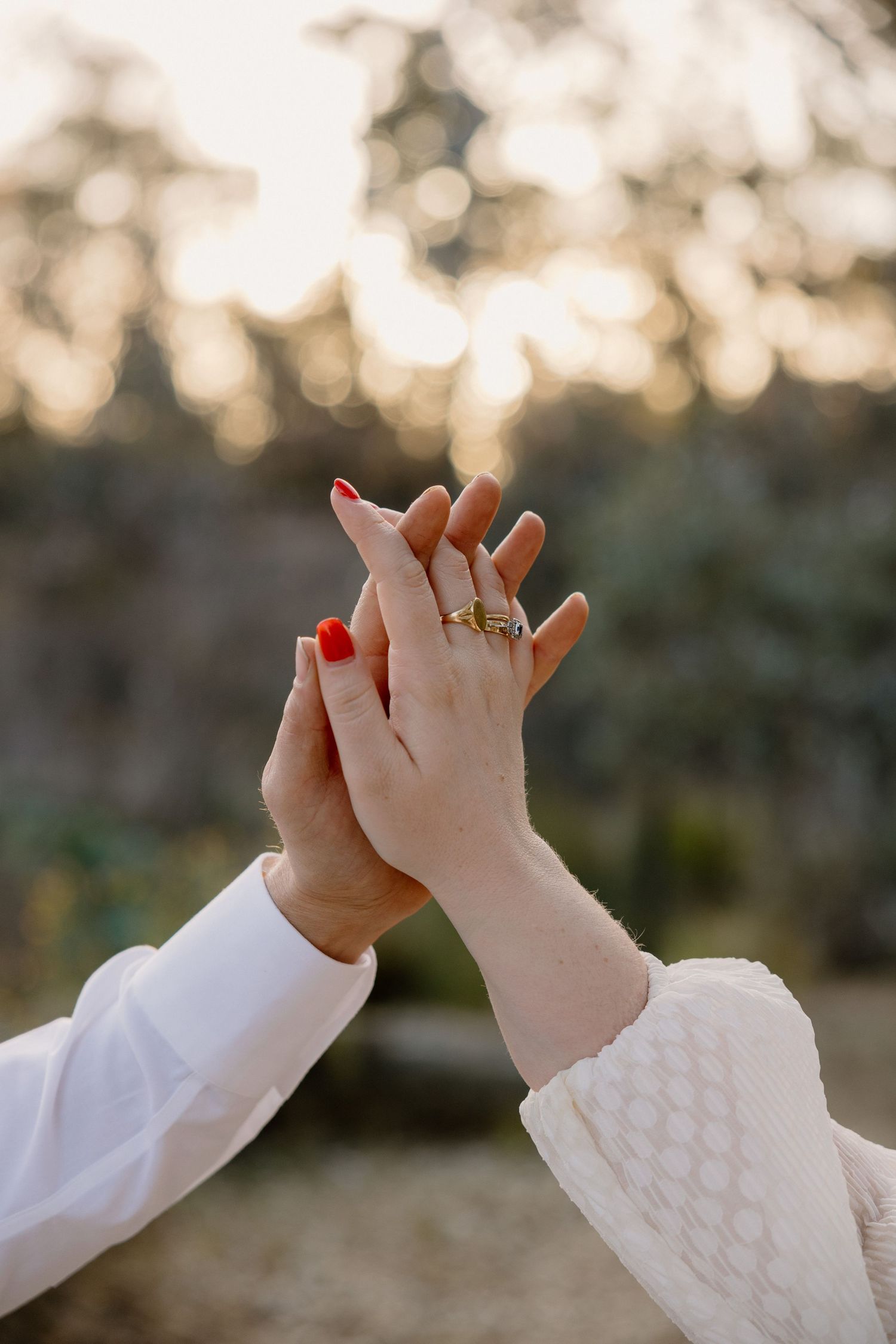 Two hands touch in tender moment outdoors with engagement ring visible against soft bokeh sunset background.