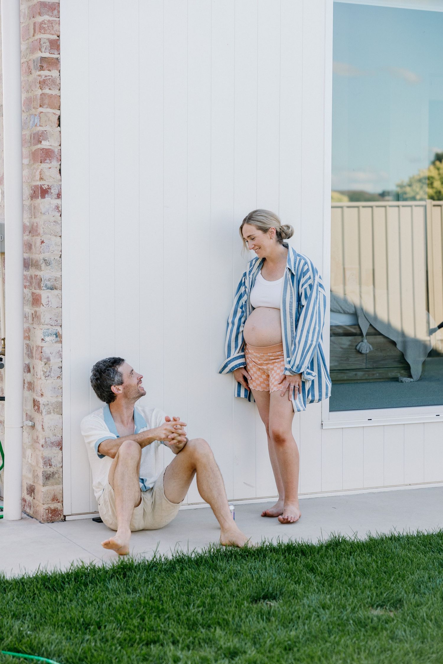 A couple sitting and interacting playfully on a porch with a grassy lawn in front of a brick wall.