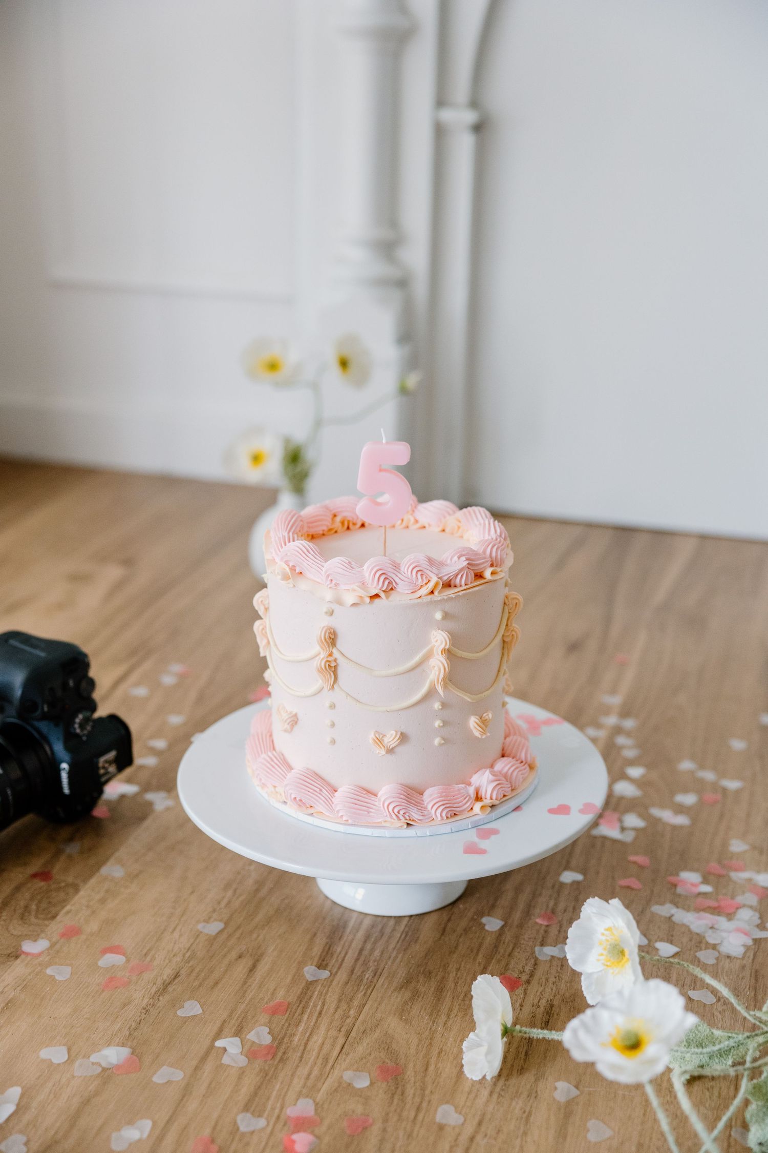 A delicate pink and white buttercream cake decorated with ruffles sits on a white cake stand with scattered flowers nearby.