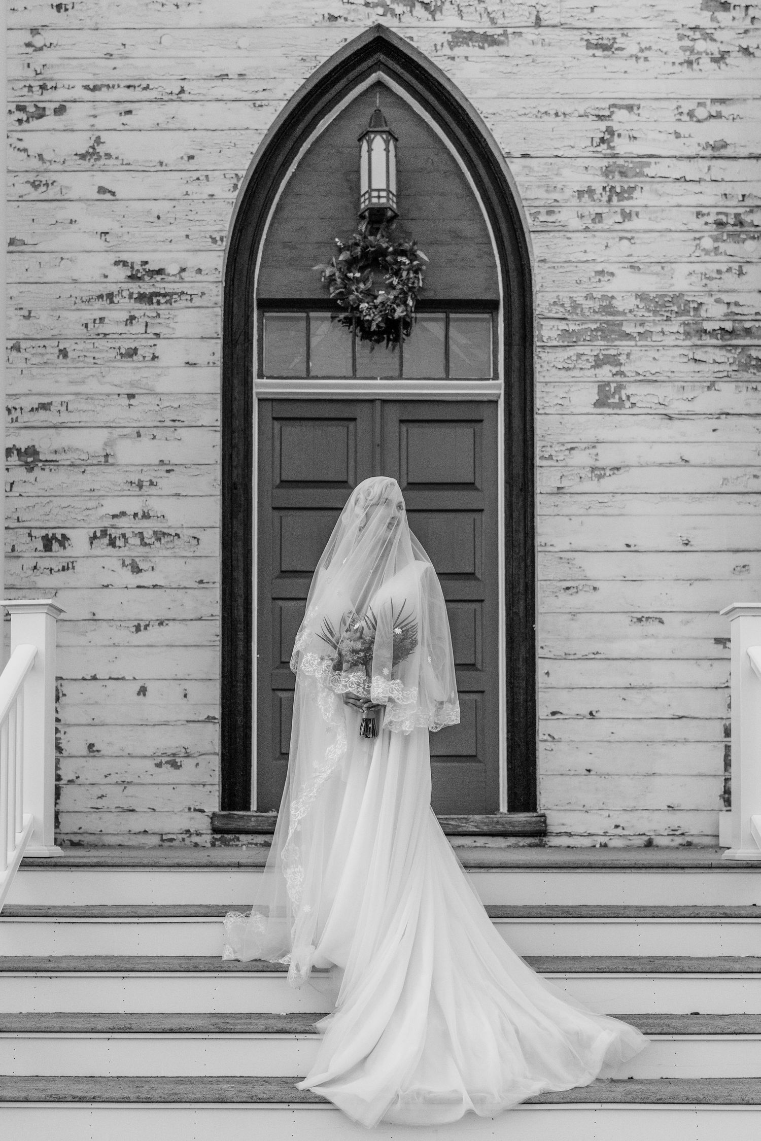 Bride in white gown and veil stands on church steps before gothic arched door in black and white photo.