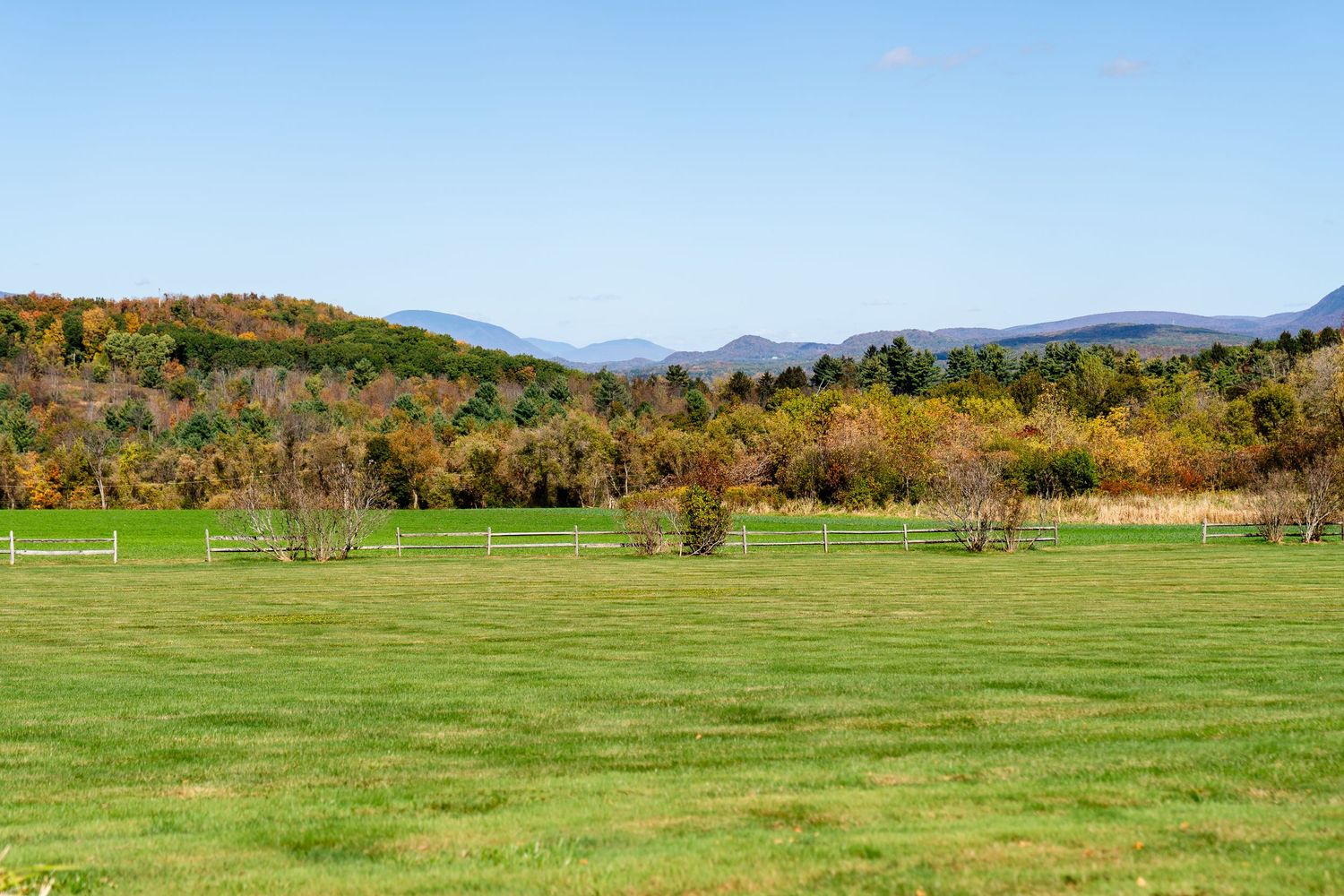 Lush green farm fields with white fence, colorful autumn trees, and distant blue mountains under a clear sky.
