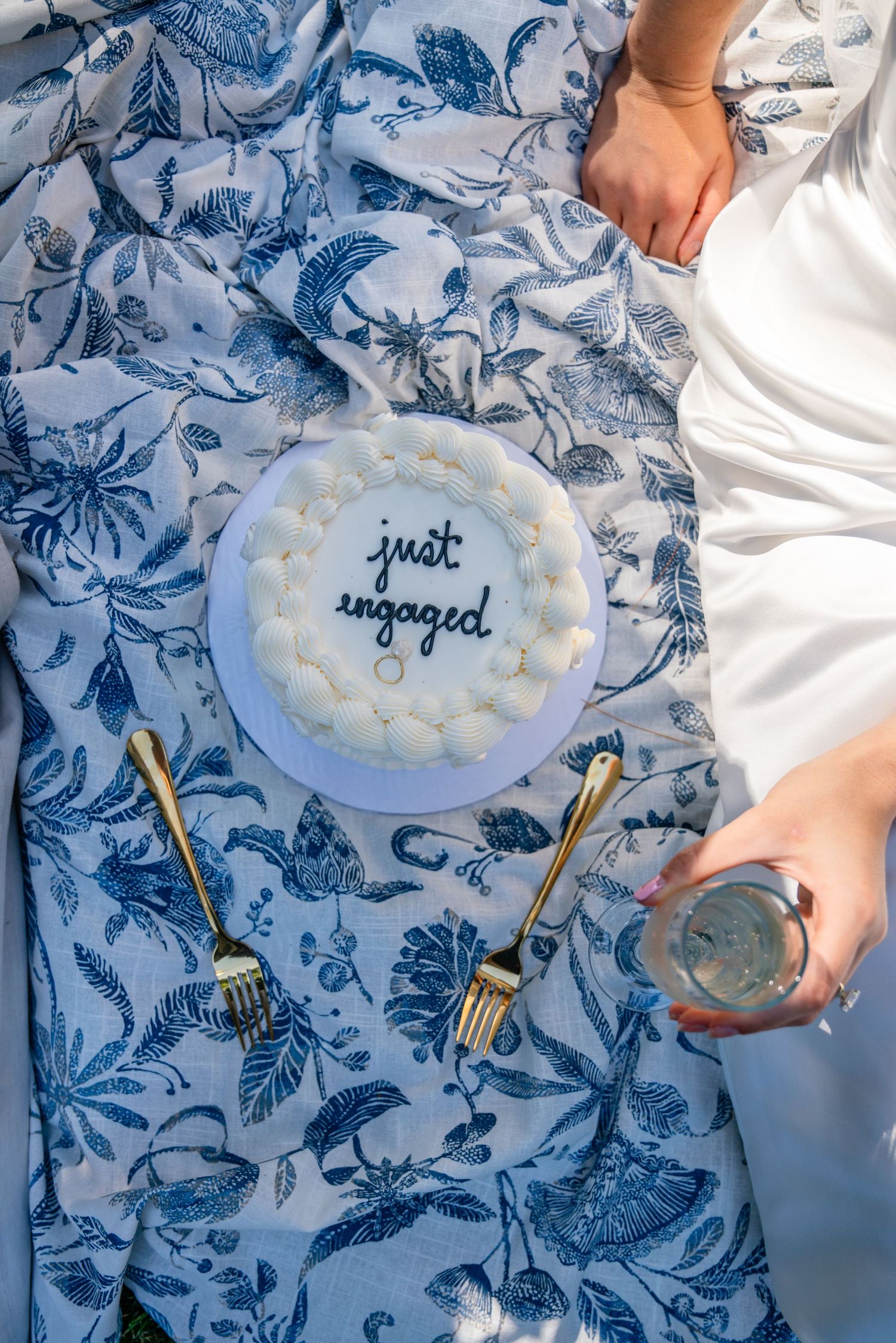 A blue and white 'Just Engaged' cake on floral patterned fabric with champagne glasses nearby.