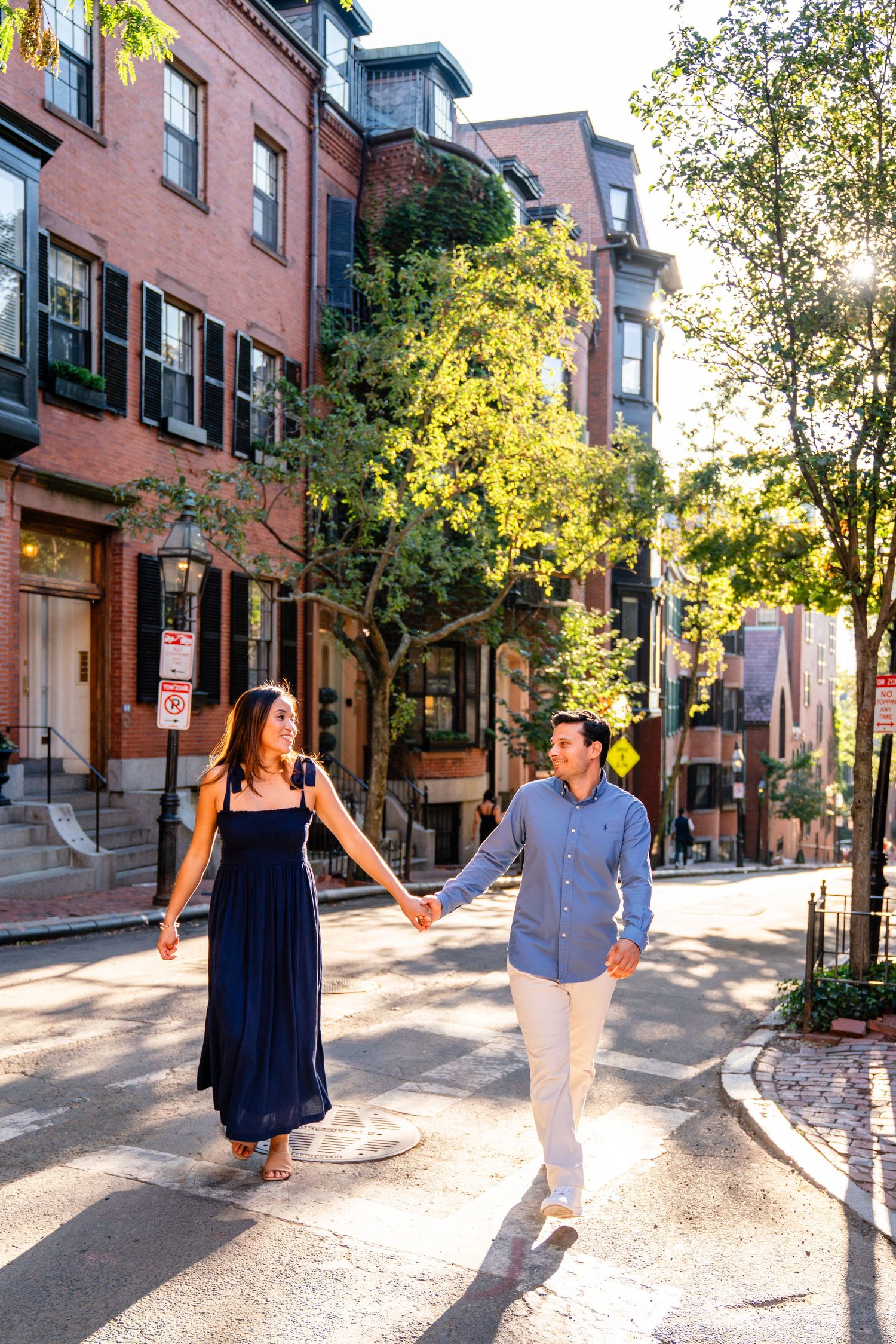 Couple holding hands walking on a sunny Boston street lined with brick brownstones and green trees.