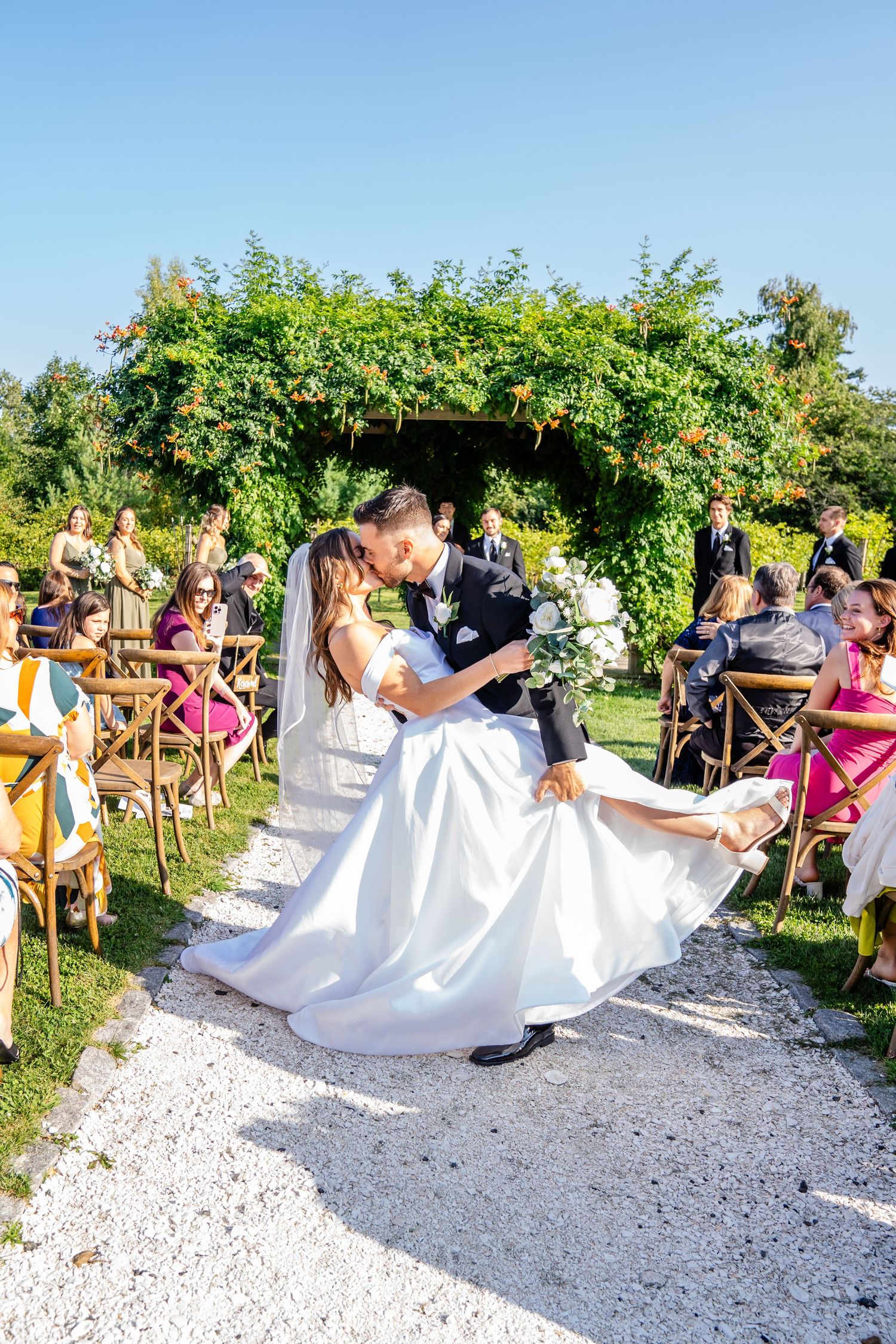 Groom dips bride for a kiss at outdoor wedding ceremony with guests seated in wooden chairs under green arch.