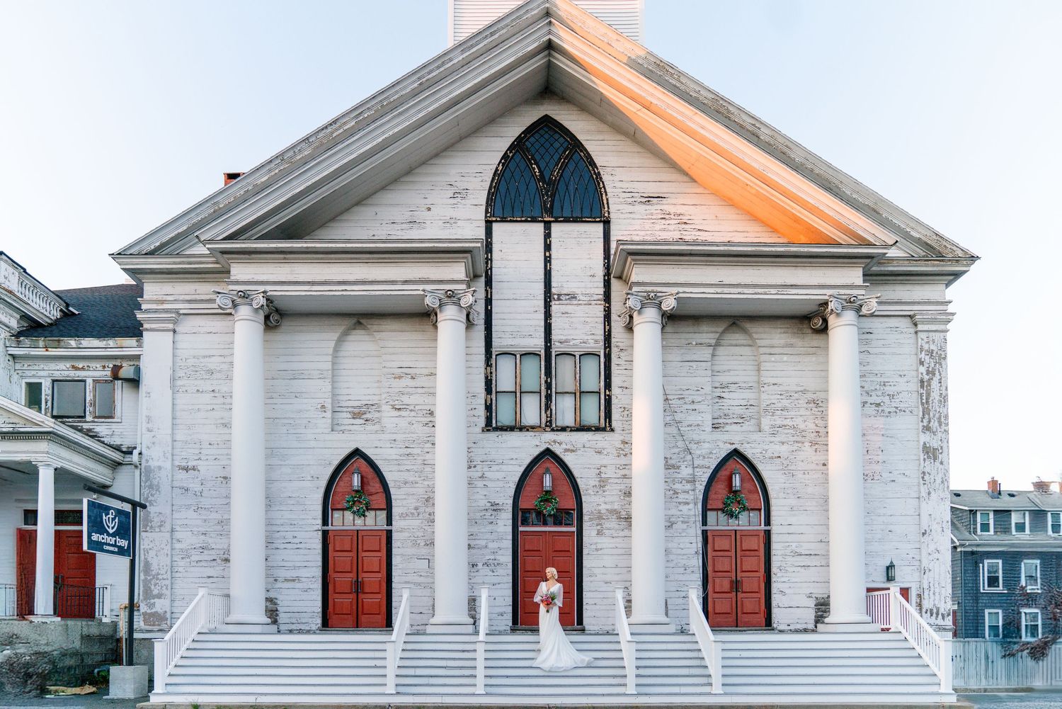 Bride standing on steps of a white historic church with red arched doors during a wedding ceremony.