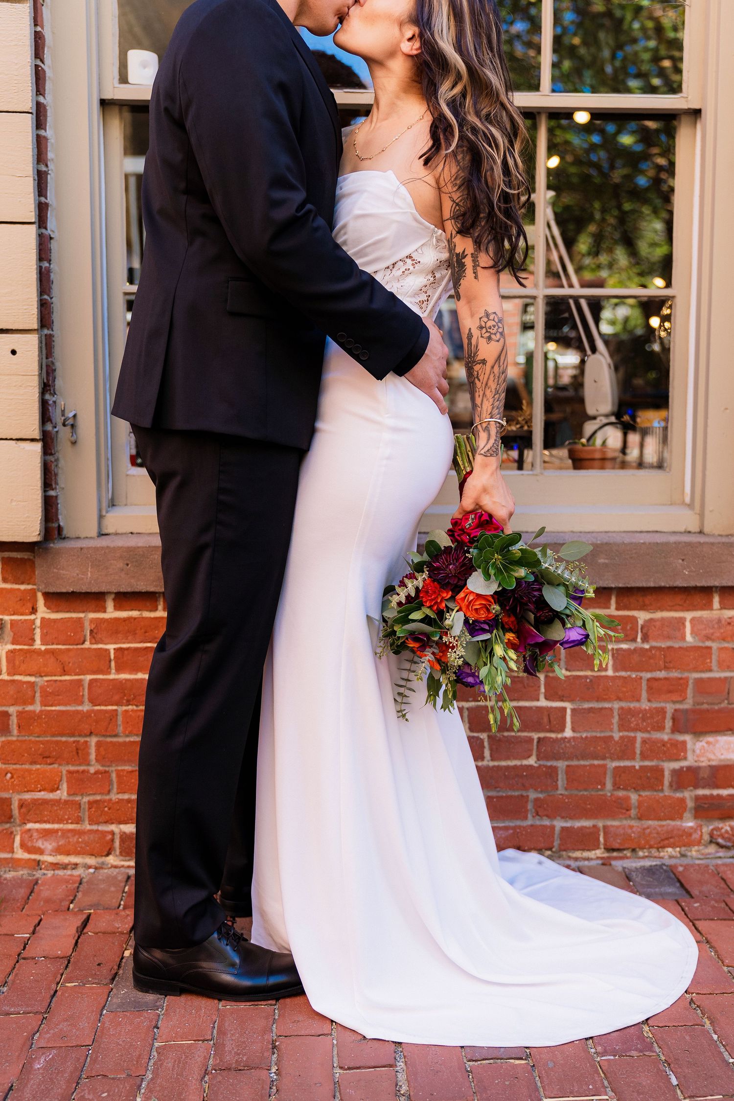 Bride and groom sharing a kiss in front of a brick wall, bride holding a vibrant red and purple wedding bouquet.