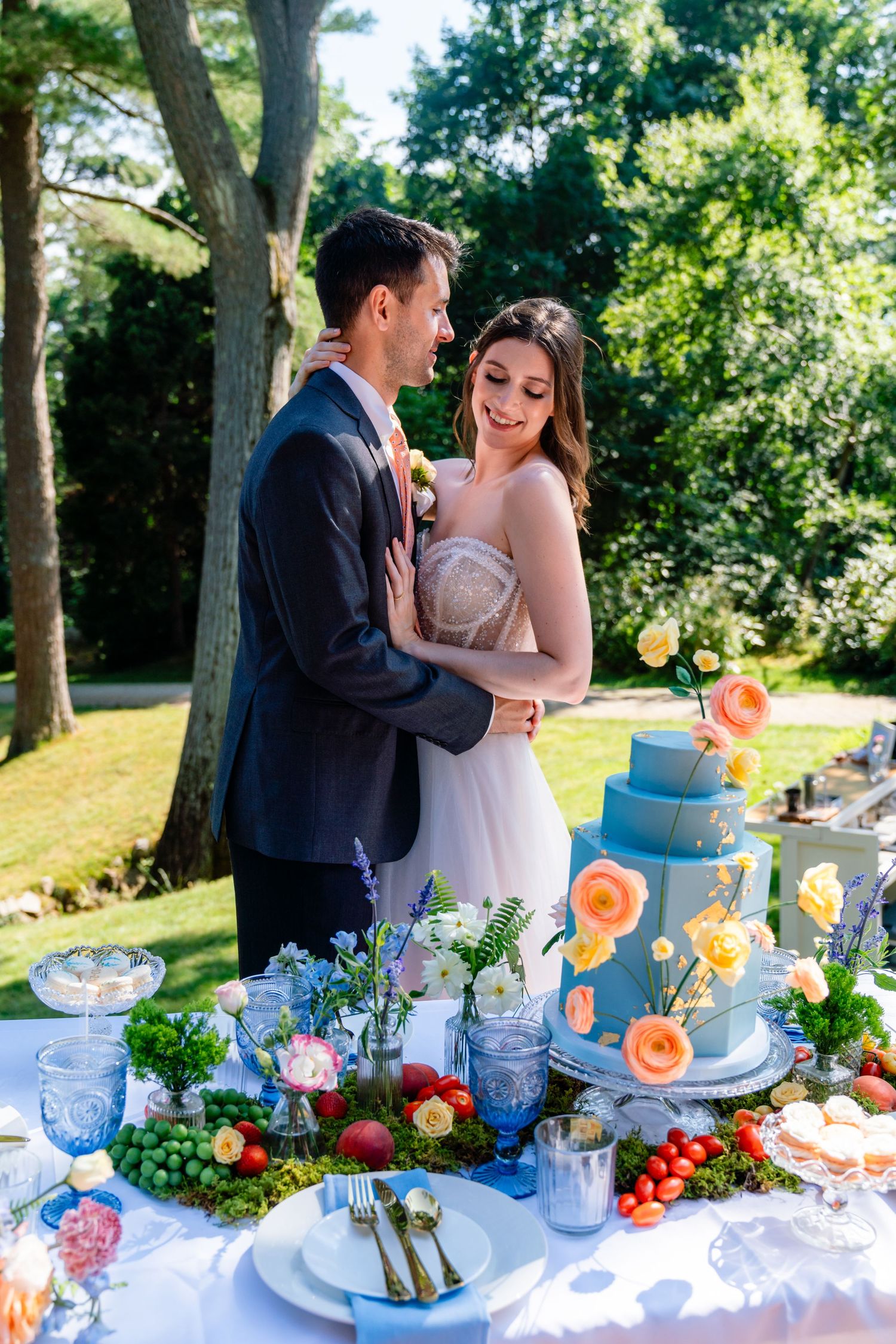 Couple embracing at outdoor wedding reception table with blue tiered cake and colorful floral arrangements in garden setting.