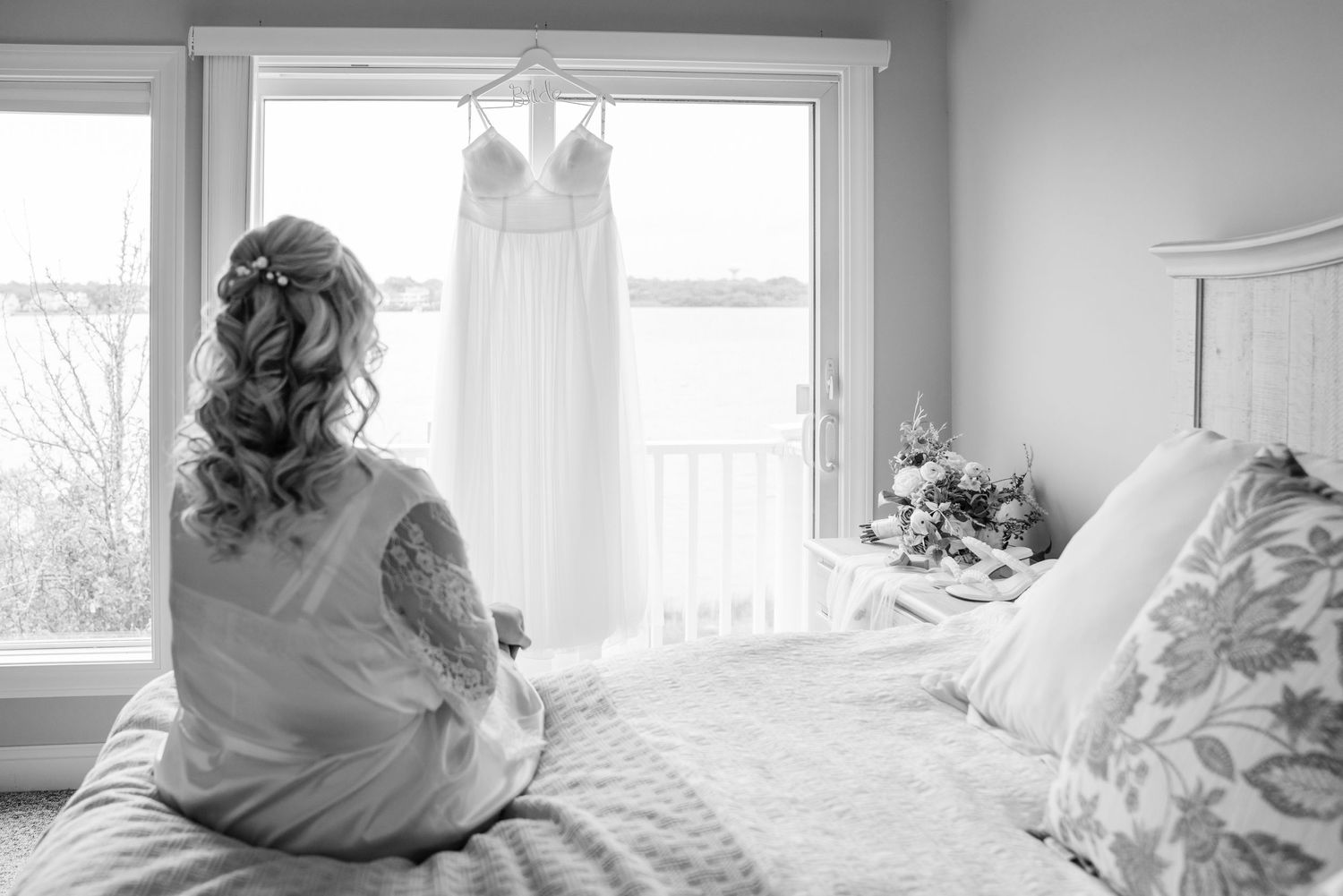 Wedding dress hanging in window while someone sits on bed looking at it in black and white photo.