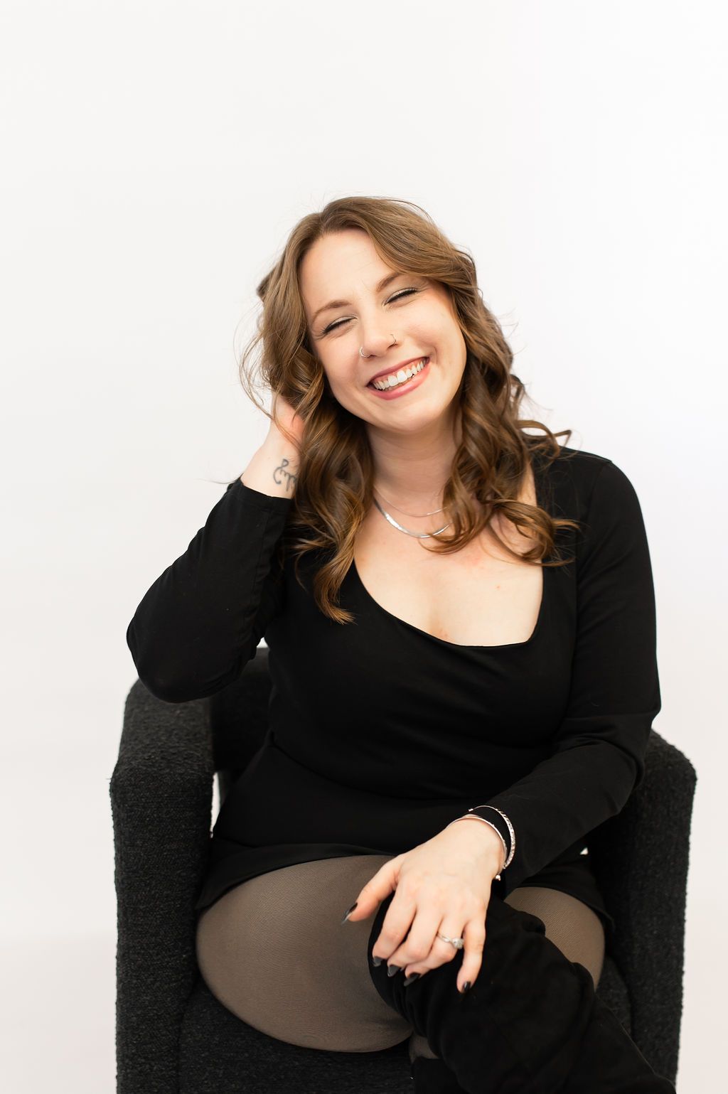 A smiling young woman with wavy brown hair sits in a chair wearing a black outfit against a white background.