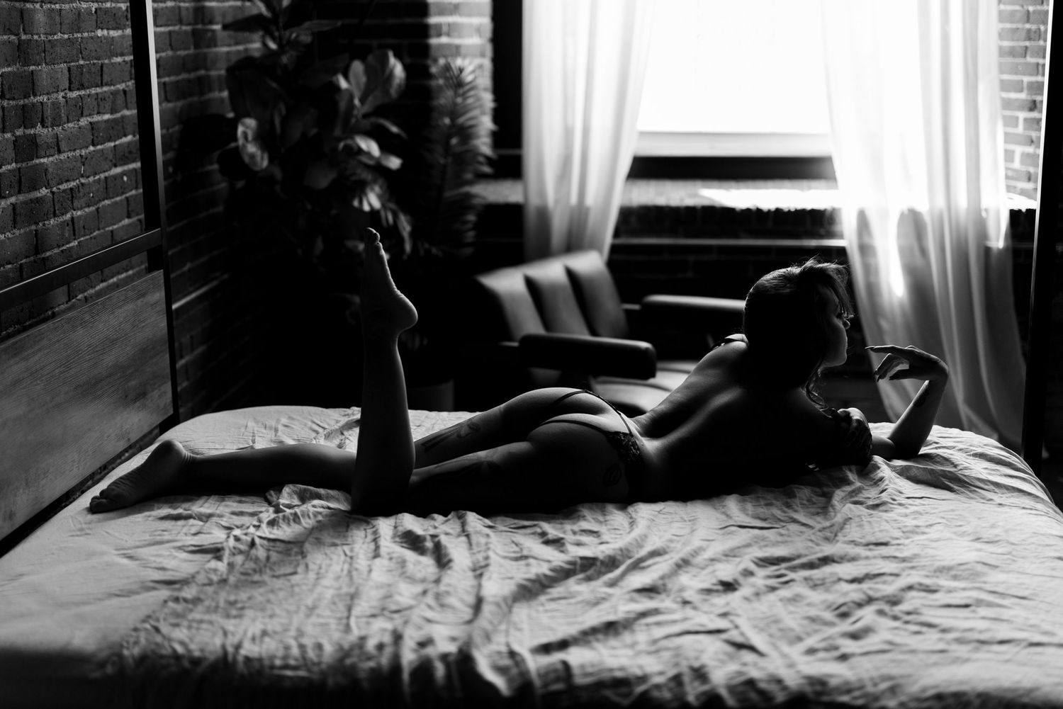 Black and white boudoir photo of a woman reclining on a bed near a sunlit window in a moody room.