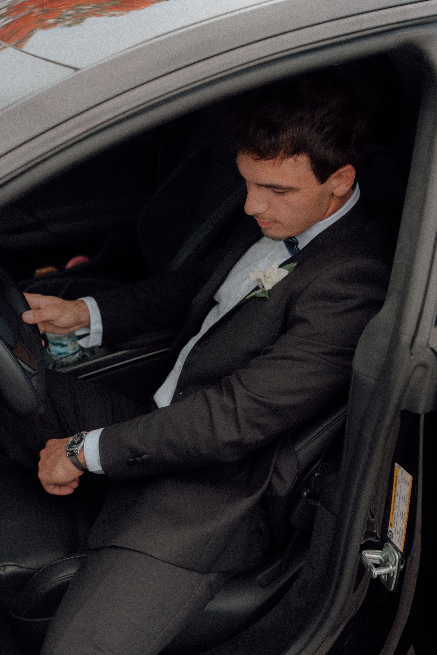 Groom in dark suit with boutonniere sitting in car, checking watch before wedding ceremony.