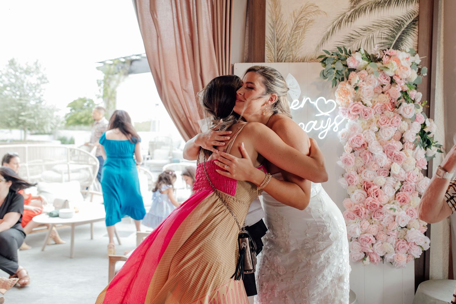Bride in white lace dress shares a warm hug with a guest at a floral backdrop.