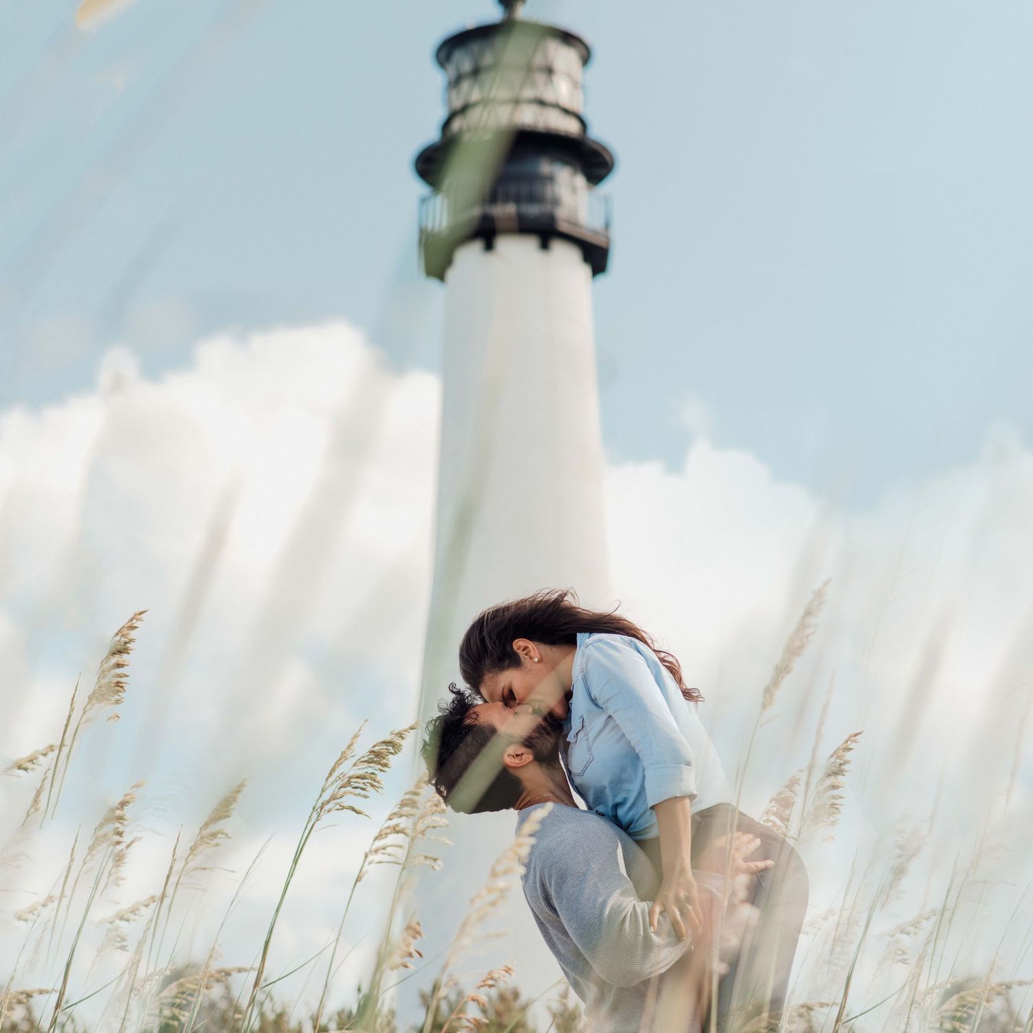 Romantic couple embracing in tall grass with a white lighthouse and cloudy sky in the background.