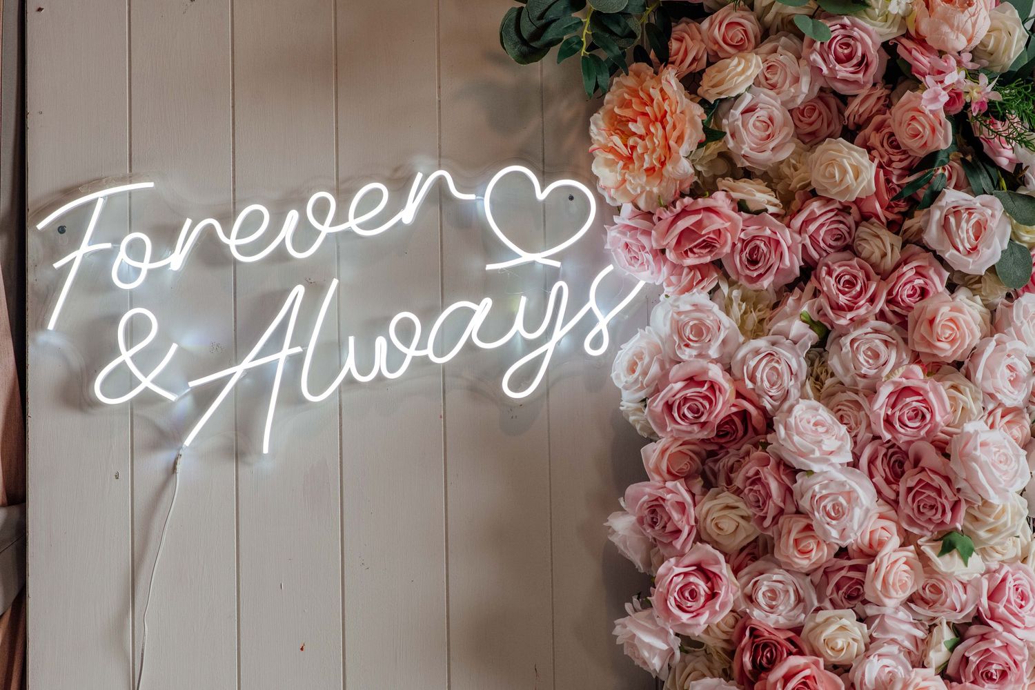 White neon Forever & Always sign with heart beside a pink rose flower wall decoration.