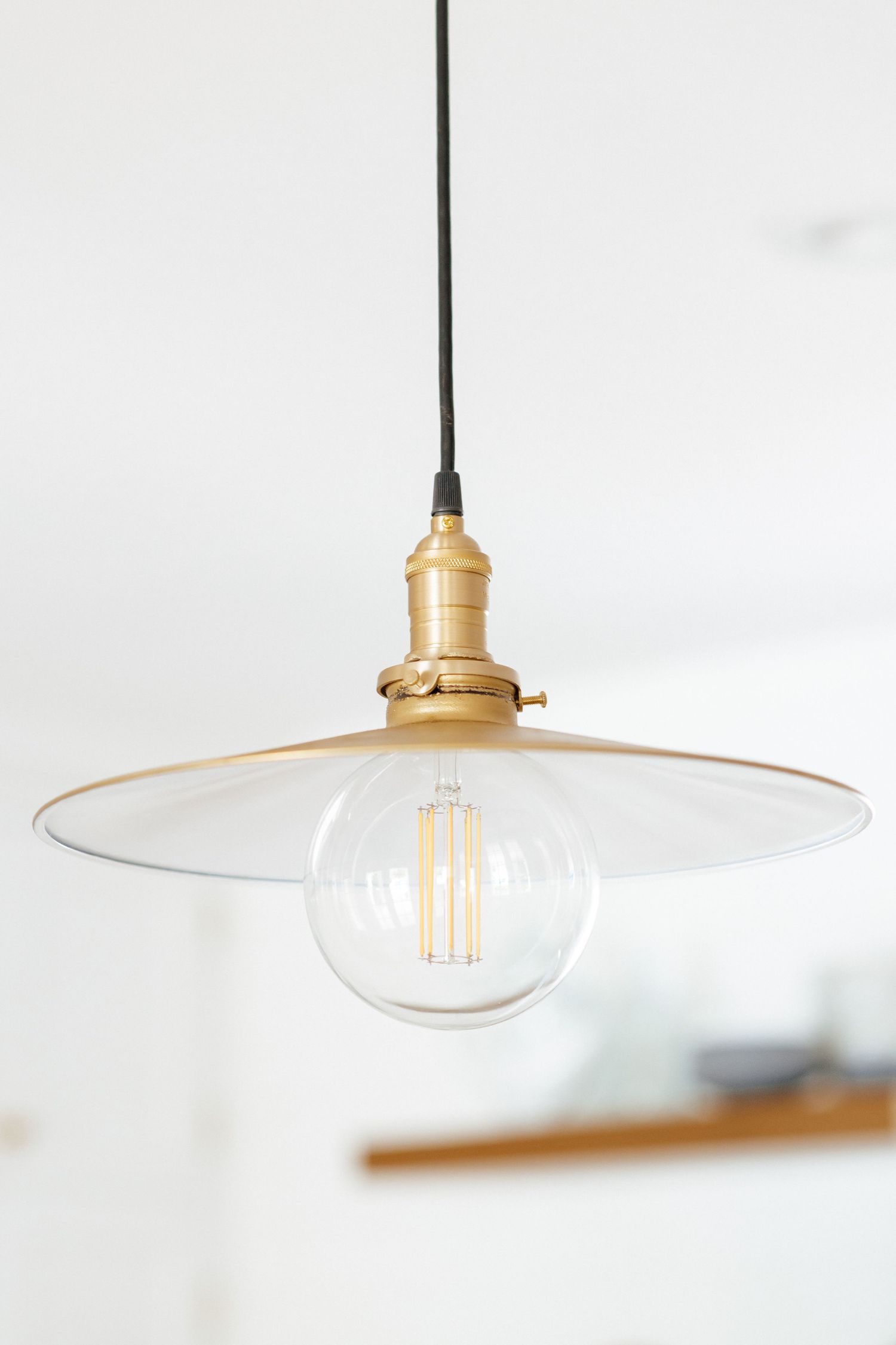Vintage brass pendant light with clear glass globe Edison bulb and flat disc shade hanging from black cord.