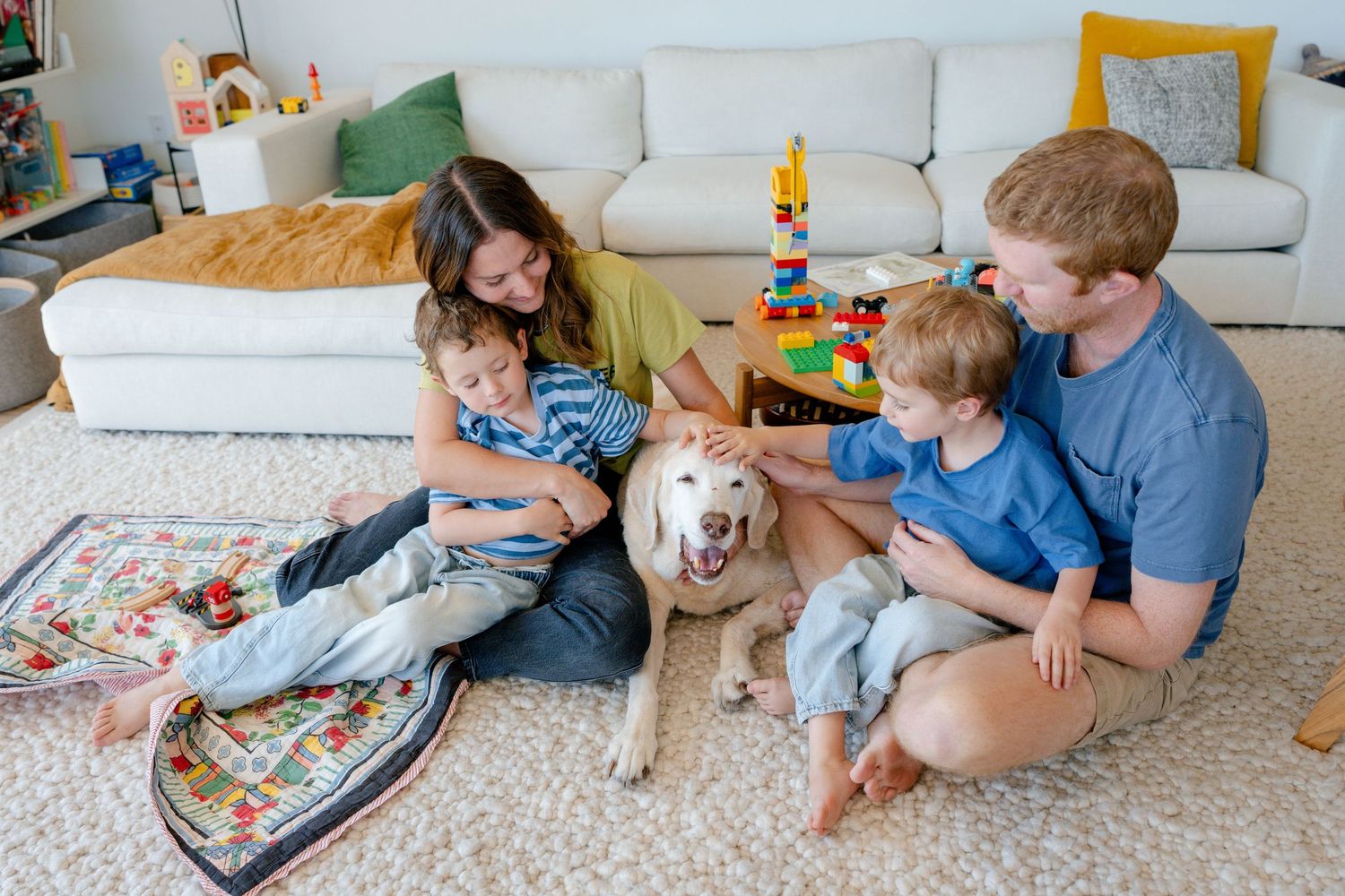 Family of four with two young boys and a golden dog sitting together on a cozy living room floor.