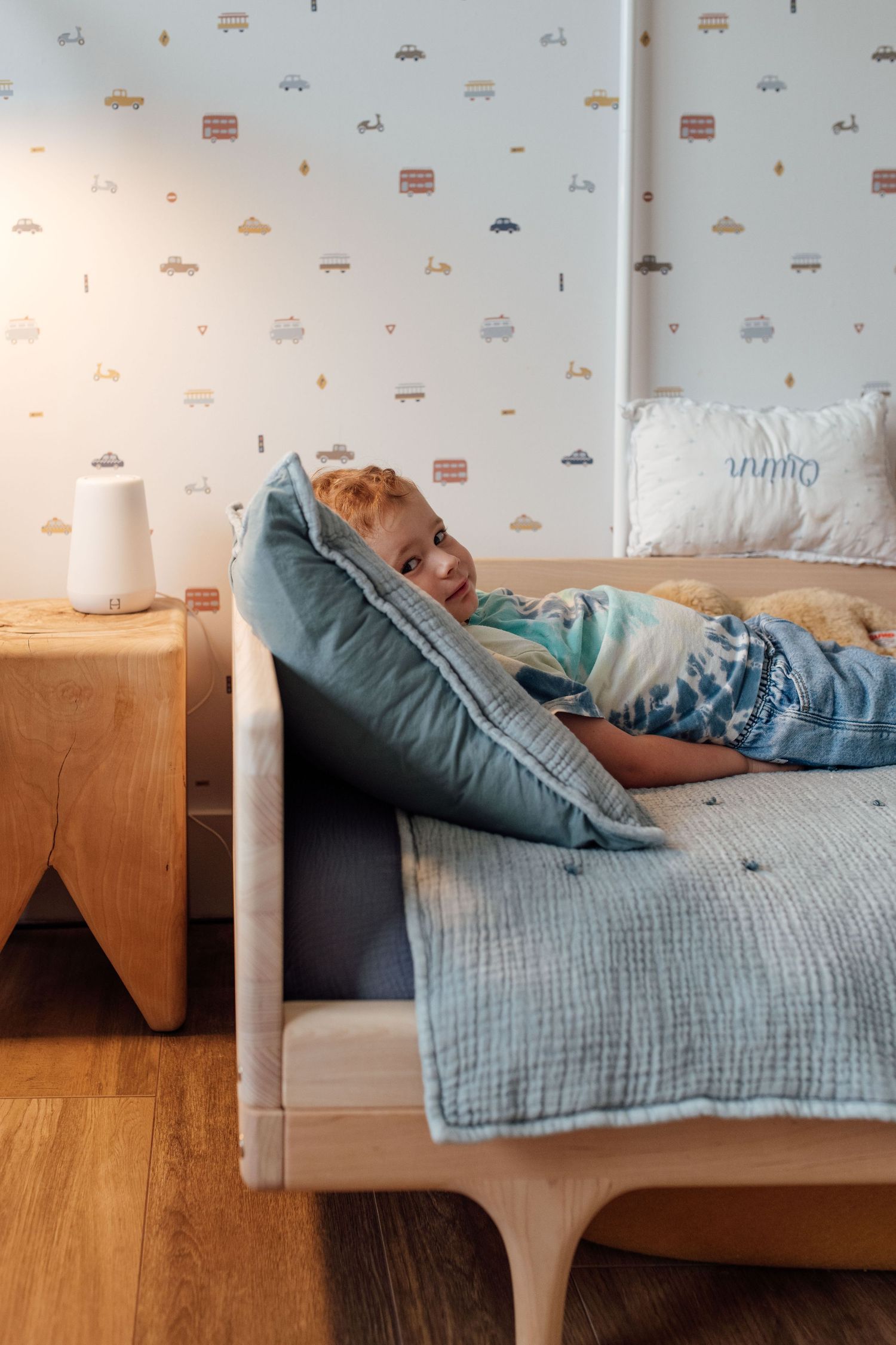 Young child lying on a tufted bed with blue pillow in a kids room with vehicle patterned wallpaper.